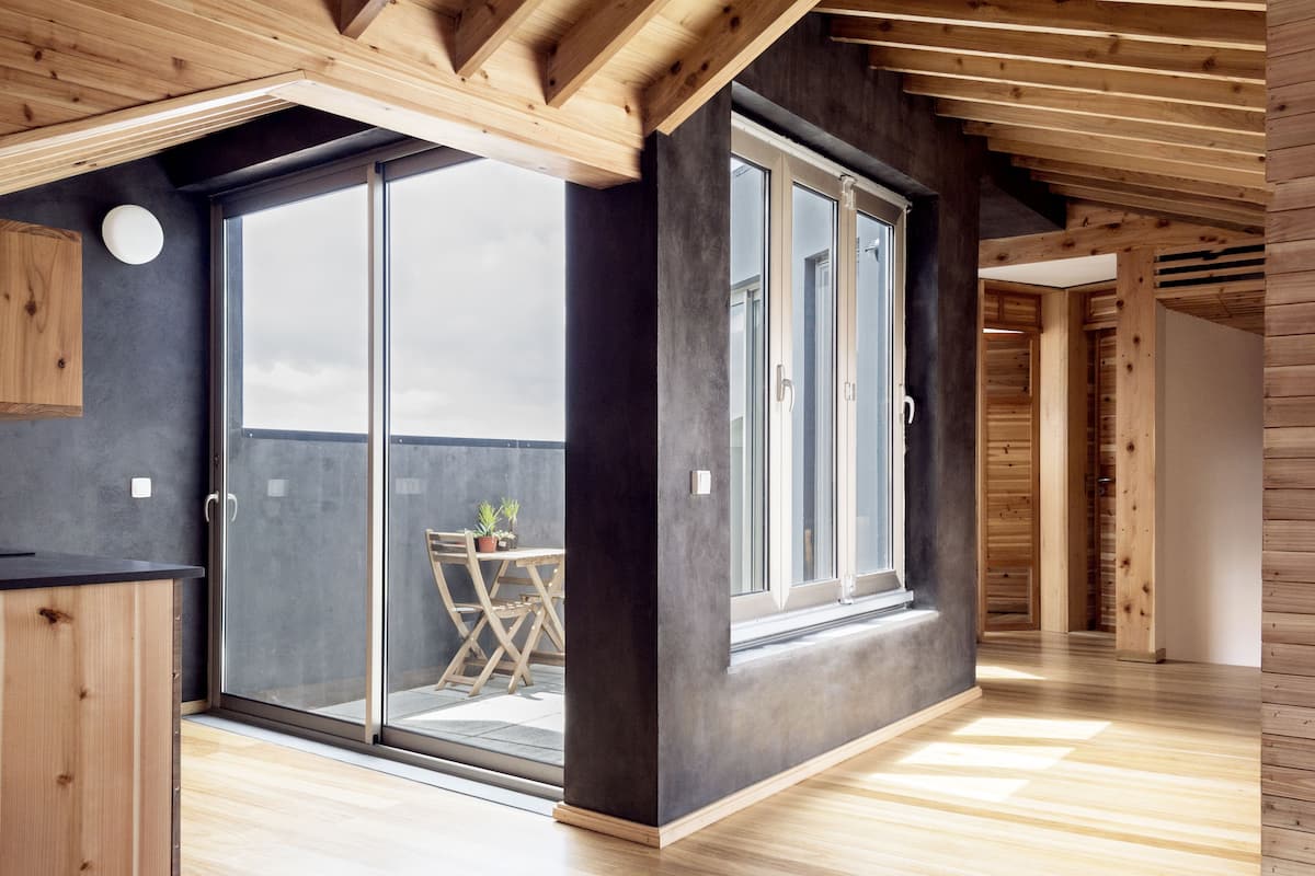 A modern interior with a large sliding glass door leading to a balcony, wooden beams on the ceiling, and a dark wall.