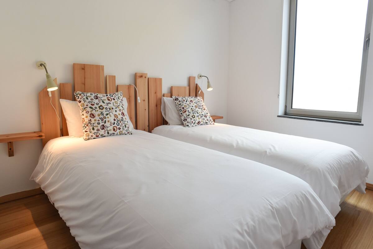 A room with two beds, each with a wooden headboard and a white bedspread. There are two bedside lamps and a window providing natural light.
