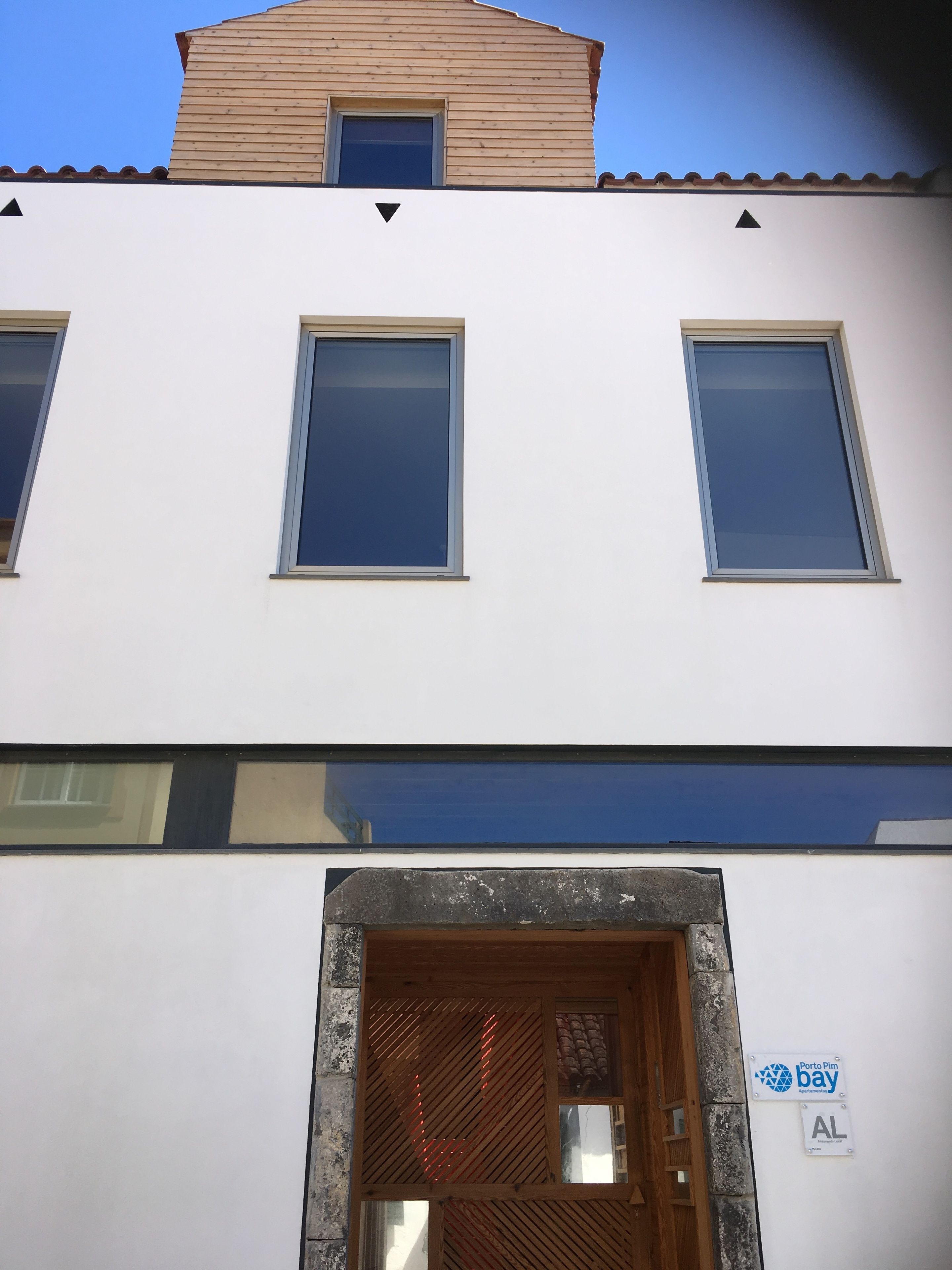 A white building with three windows and a wooden door. There is a sign on the door that reads "Porto Pim bay".