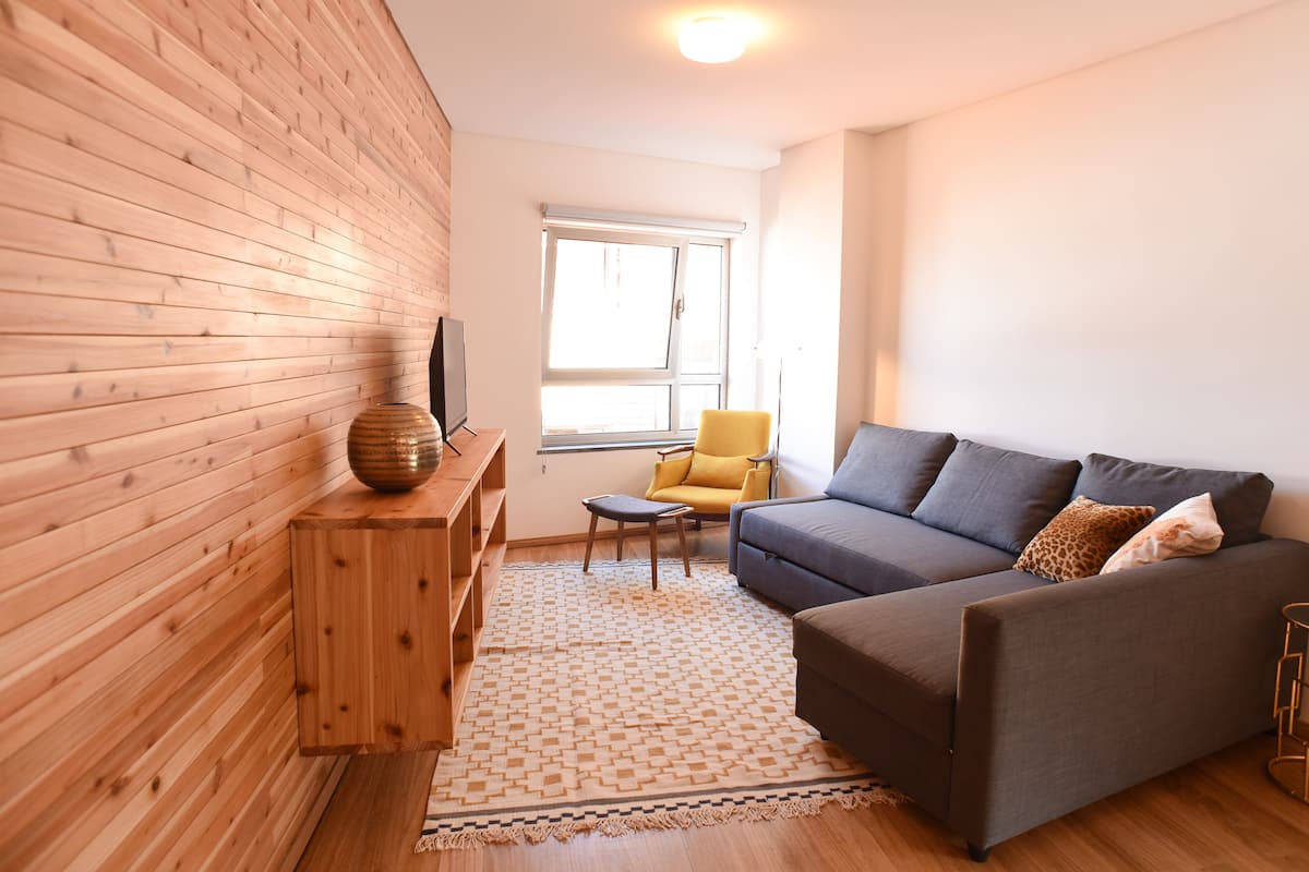 A modern living room with a wooden wall, a grey sofa, a yellow armchair, and a patterned rug.