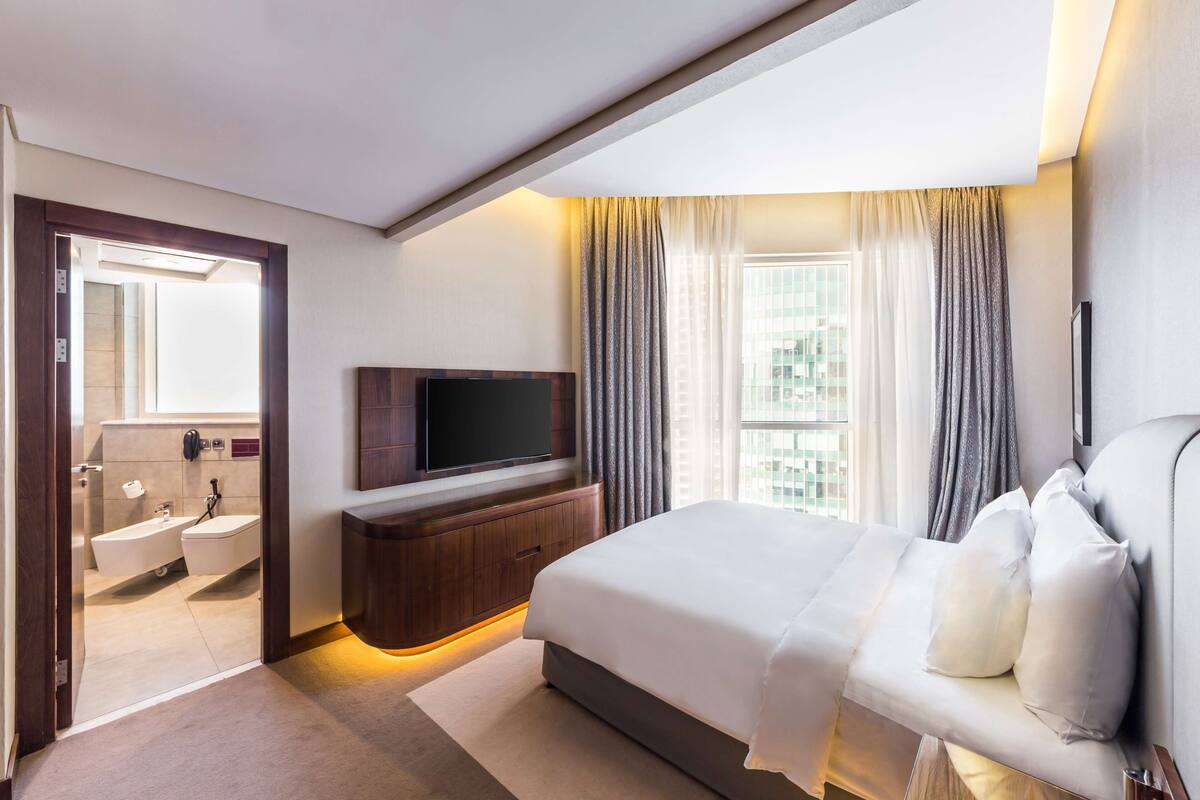 Ein modernes Hotelzimmer mit einem großen Bett, einem an der Wand montierten Fernseher, einem Badezimmer mit Badewanne und Dusche sowie einem Blick auf die Stadt durch große Fenster.