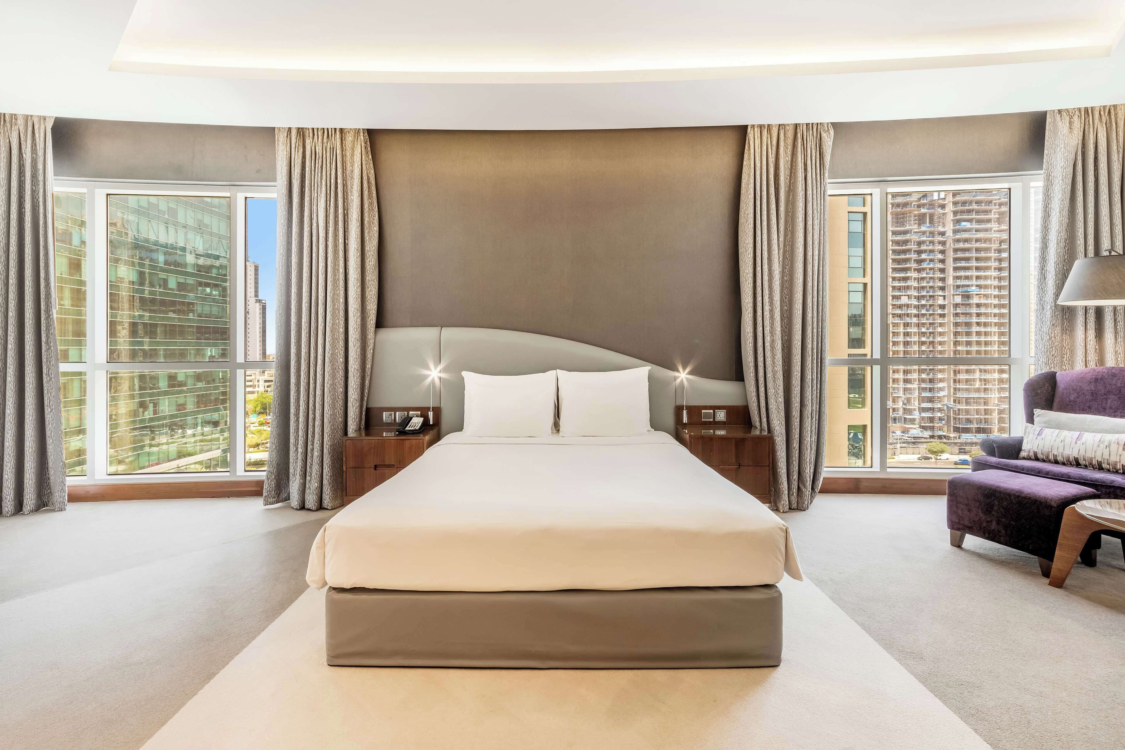 A modern hotel bedroom with a large bed, two bedside tables, a nightstand, and a view of the city through large windows.