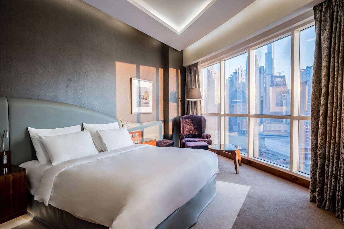 Ein modernes Hotelzimmer mit einem großen Bett, einem Nachttisch, einem Sessel und Blick auf die Stadt durch große Fenster.