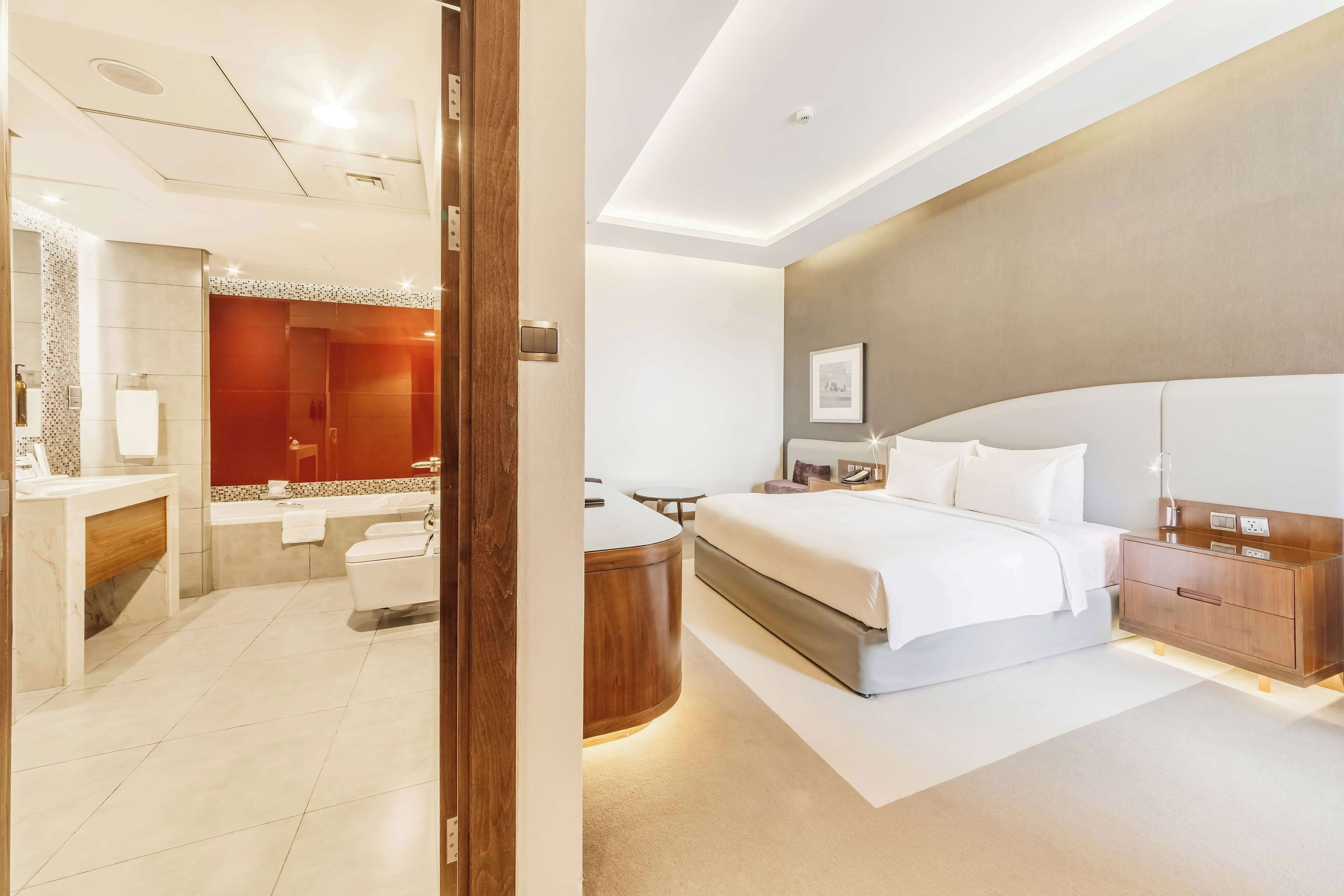 A modern hotel room with a large bed, a wooden nightstand, a bedside table, a desk, and a bathroom with a shower and toilet.