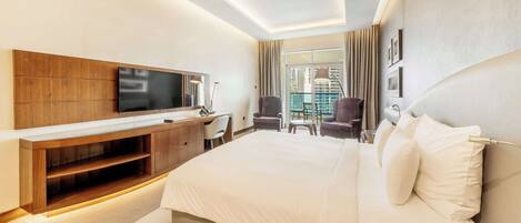 A modern hotel room with a large bed, a TV mounted on a wooden console, a desk with a chair, and a view of the city through large windows.
