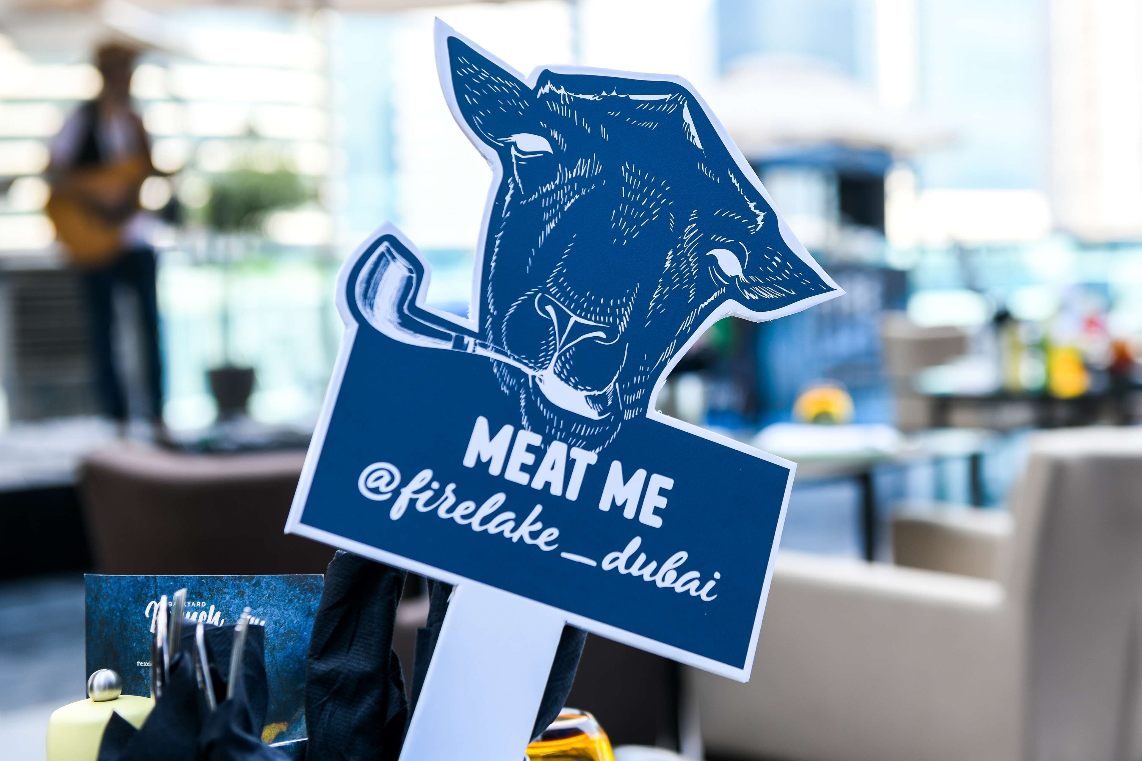 A sign with a cow illustration and the text "MEAT ME @firelake_dubai" in a modern indoor setting.