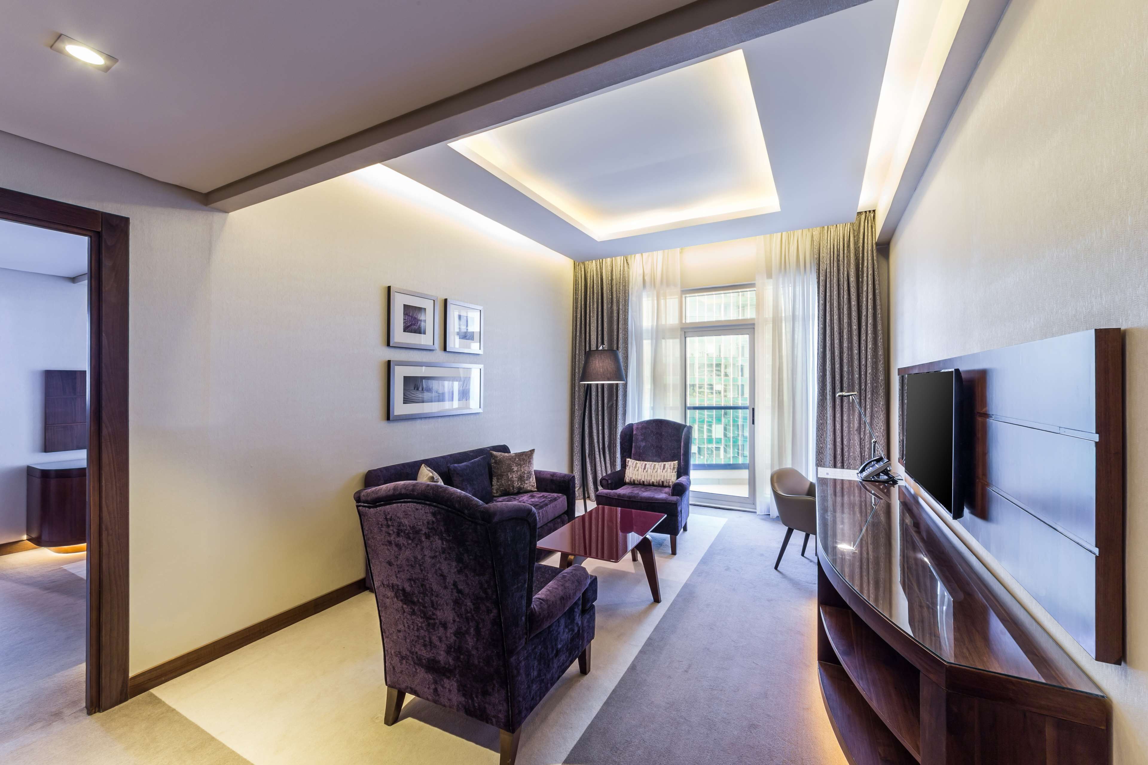 Une chambre d’hôtel moderne avec un canapé de couleur violet foncé, un fauteuil assorti, un téléviseur à écran plat et une grande fenêtre avec des rideaux.