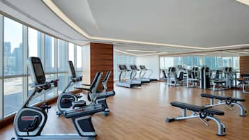 A modern gym with various cardio and strength training equipment, large windows offering city views, and a wooden floor.