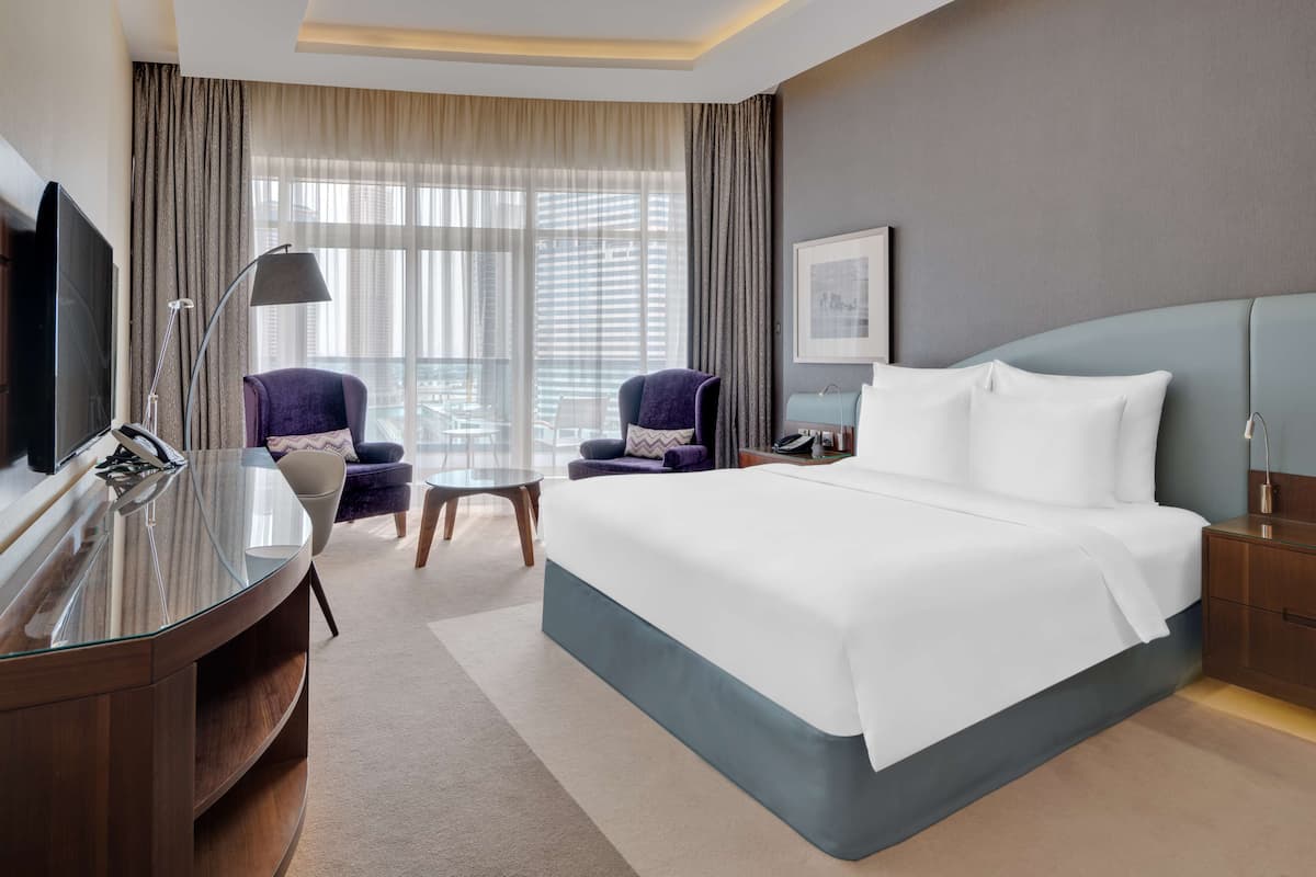 Ein modernes Hotelzimmer mit einem großen Bett, einem Schreibtisch, einem Flachbildfernseher, zwei Sesseln, einem kleinen Tisch und einem großen Fenster mit Vorhängen.
