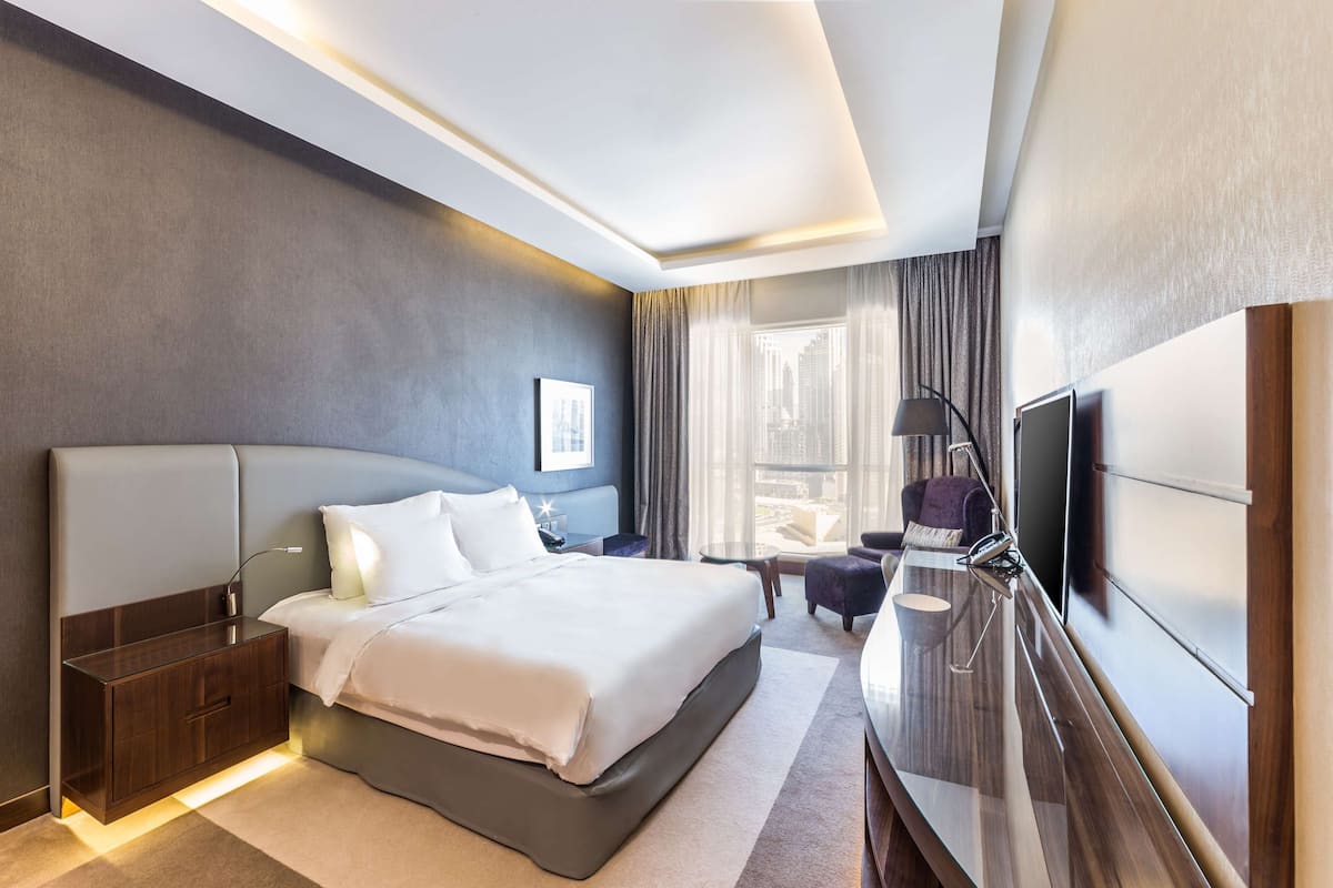 Ein modernes Hotelzimmer mit einem großen Bett, Nachttischen, einem Flachbildfernseher und Blick auf die Stadt durch große Fenster.