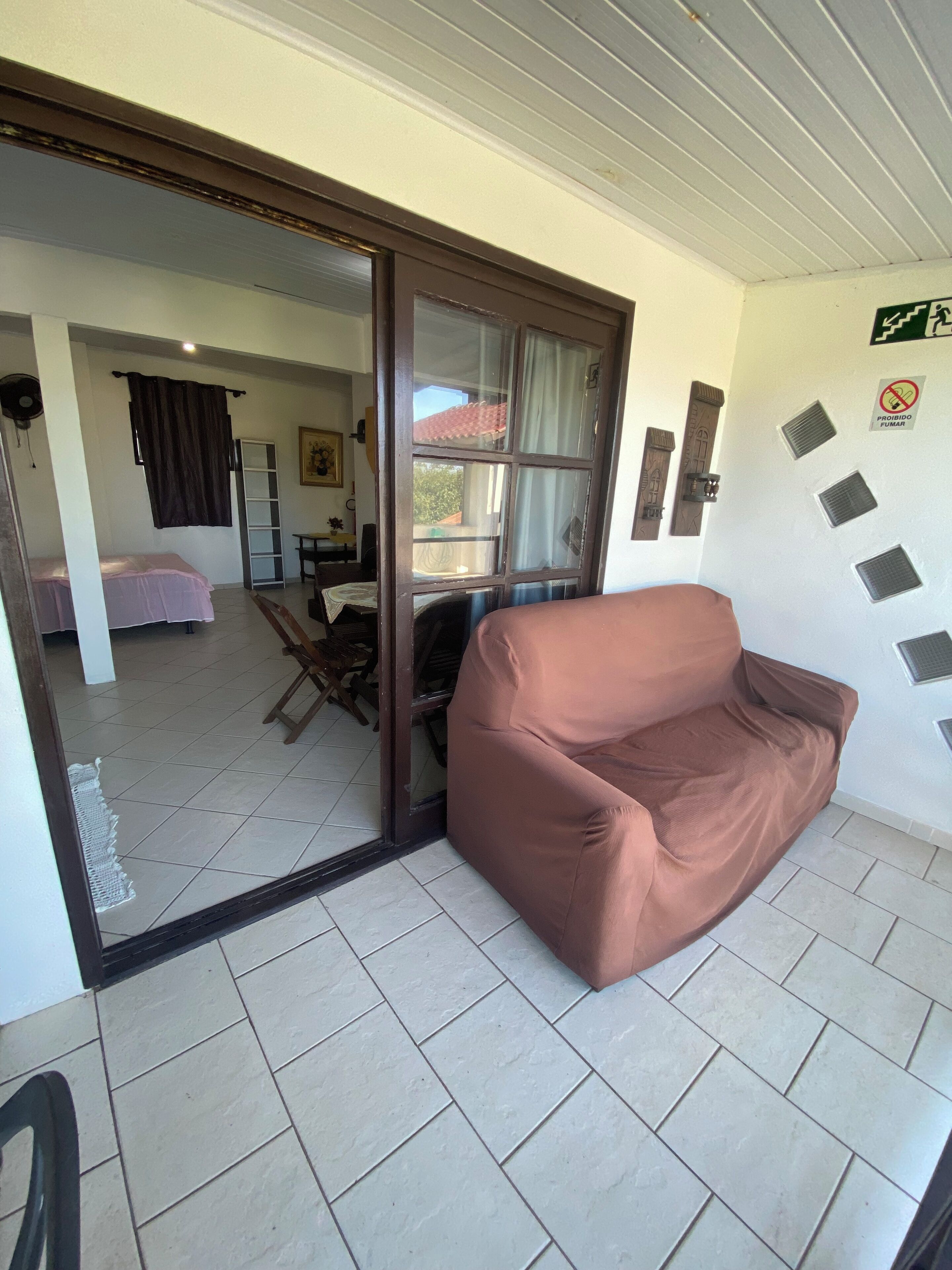 A room with a brown sofa, a sliding glass door leading to another room, tiled flooring, and a wall with ventilation grilles and a no-smoking sign.
