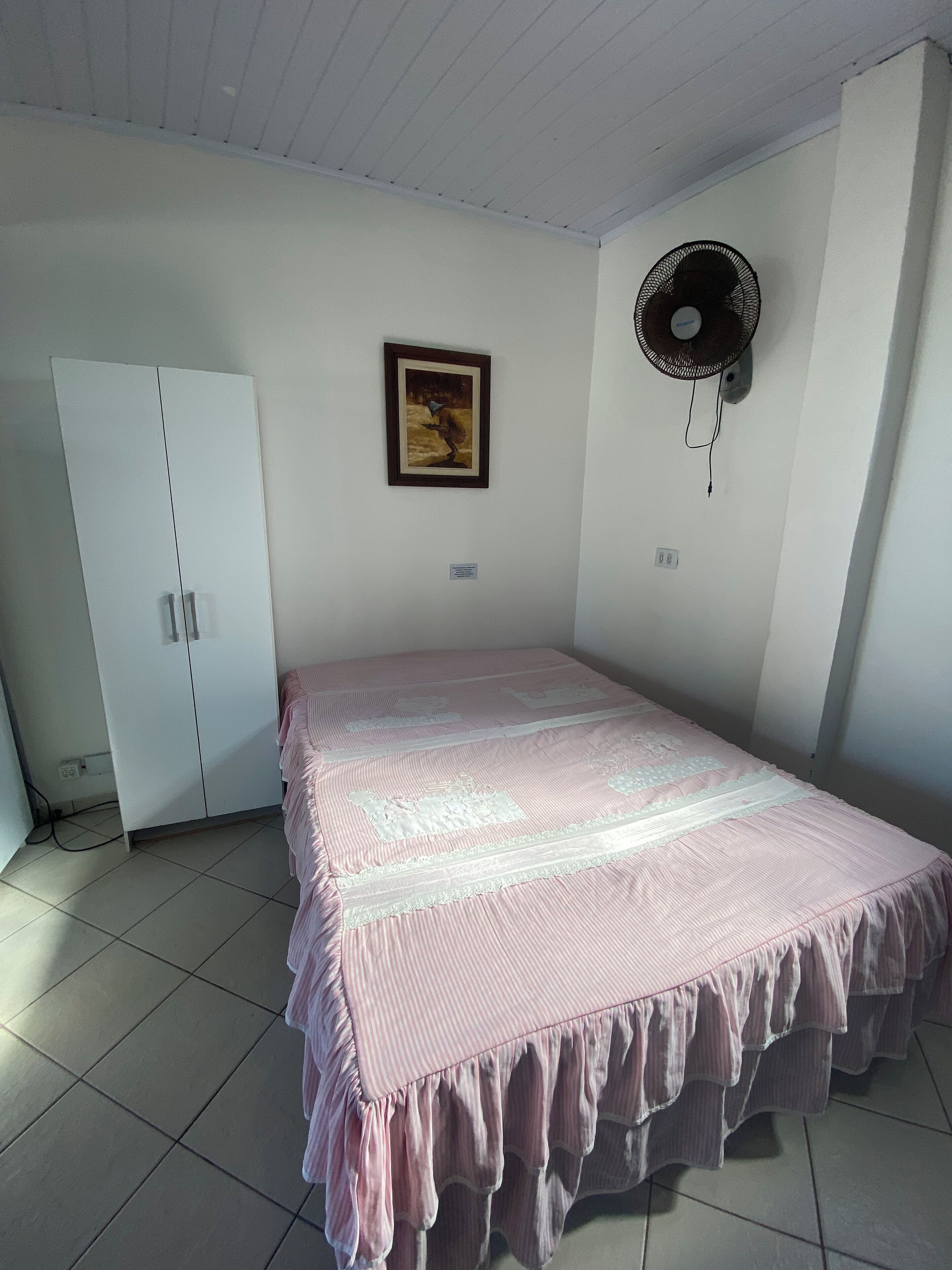 Un dormitorio con cama, armario, ventilador de pared y un cuadro enmarcado.