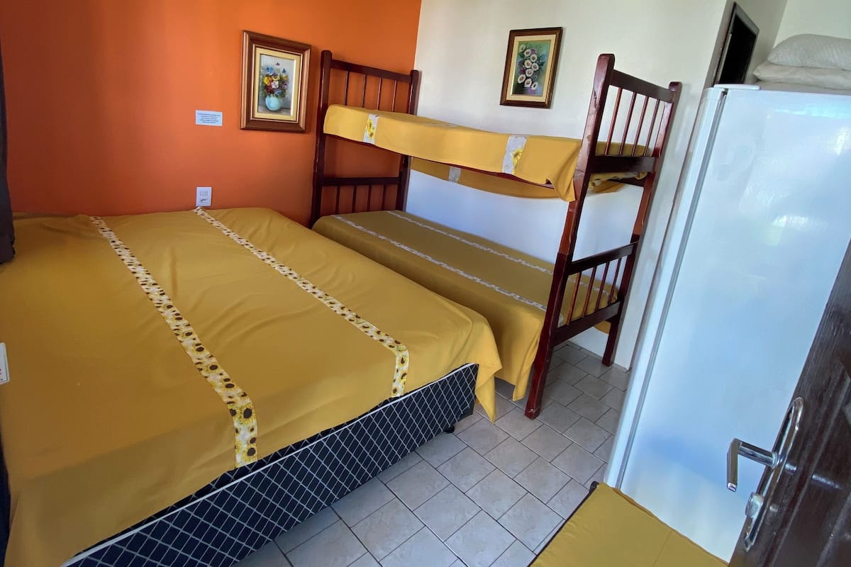 Ein Zimmer mit einem Etagenbett, einem Einzelbett, einem Sessel, einem Kühlschrank und zwei gerahmten Bildern an der Wand.
