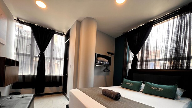 A hotel room with a large bed, two pillows with the hotel name, a bedside table, a chair, and a window with curtains.