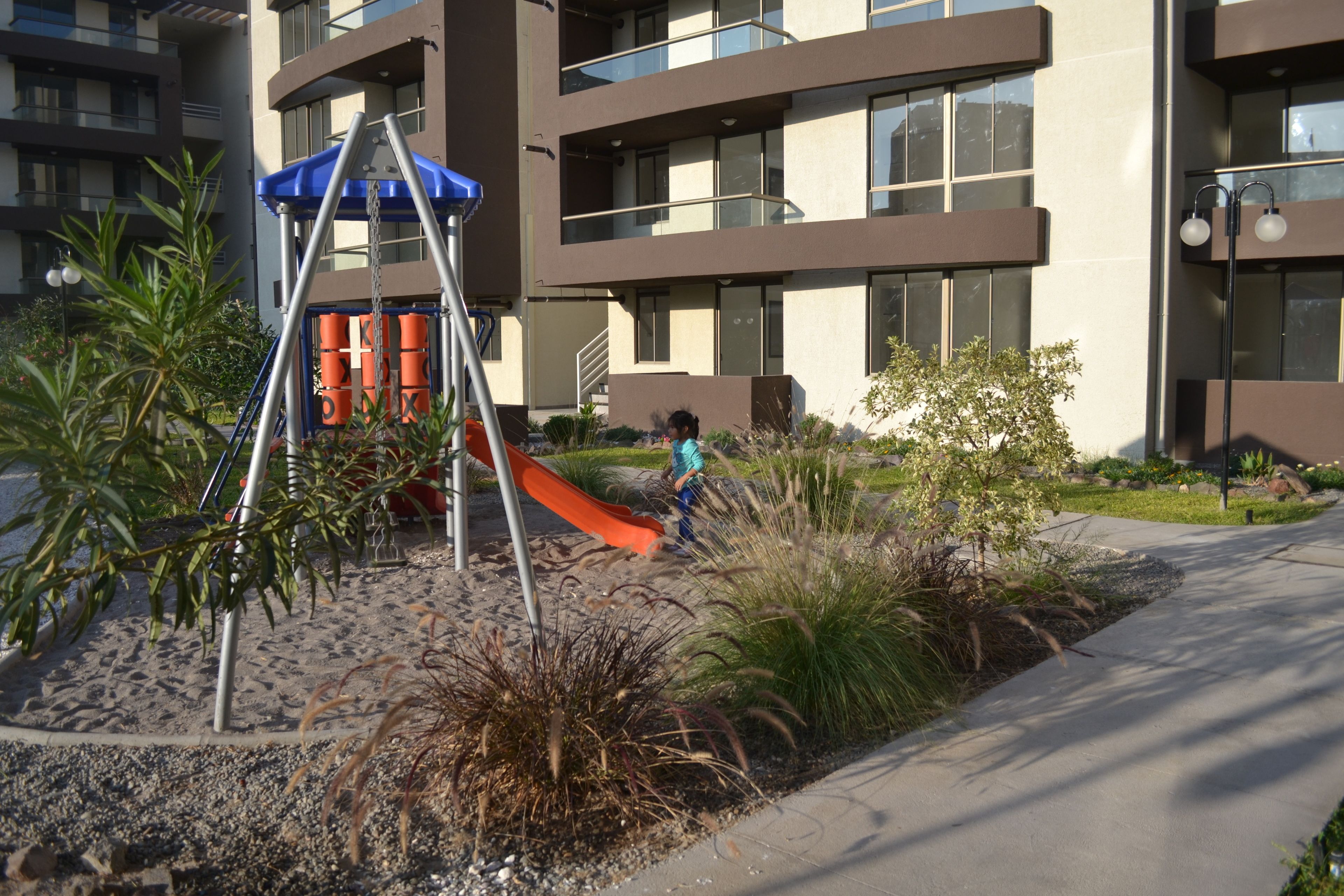 A playground with a slide and swings, surrounded by landscaped areas and a building with large windows.