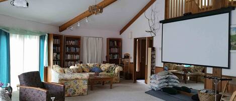 A cozy living room with a sofa, armchairs, a coffee table, and a bookshelf. A ceiling fan and projector are visible.