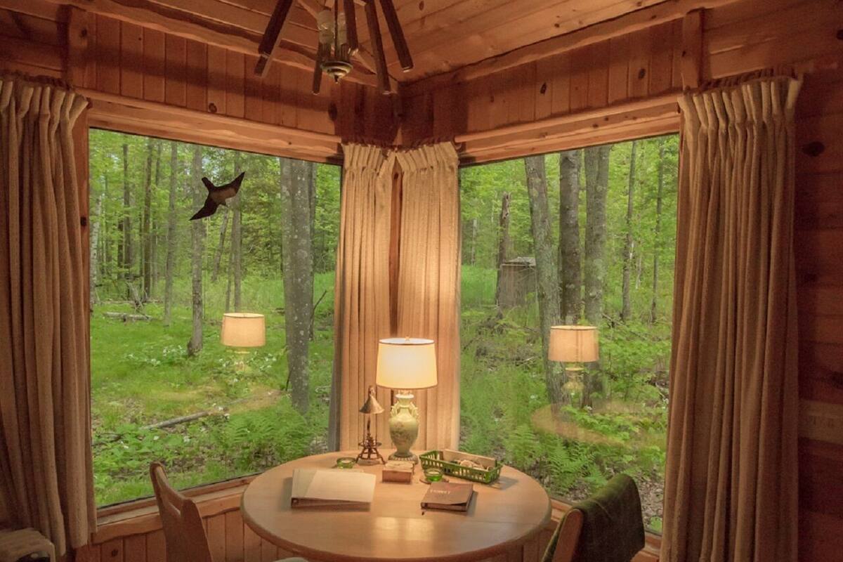 A wooden cabin interior with a round dining table, two chairs, a ceiling fan, and a window with curtains.
