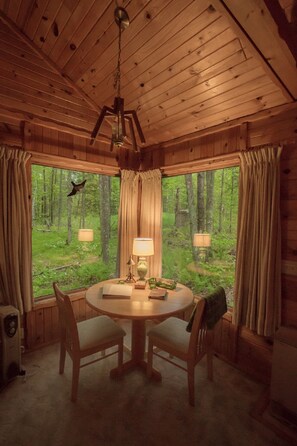 A wooden cabin interior with a round dining table, two chairs, a ceiling fan, and large windows offering a view of a forest. - Shire In The Woods (Mc Grath)