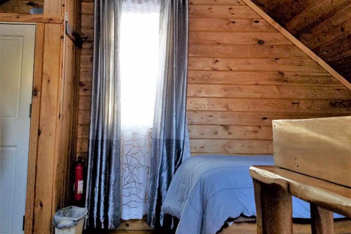 A cozy attic bedroom with wooden walls, a sloped ceiling, a bed, a bench, and a hanging light fixture.