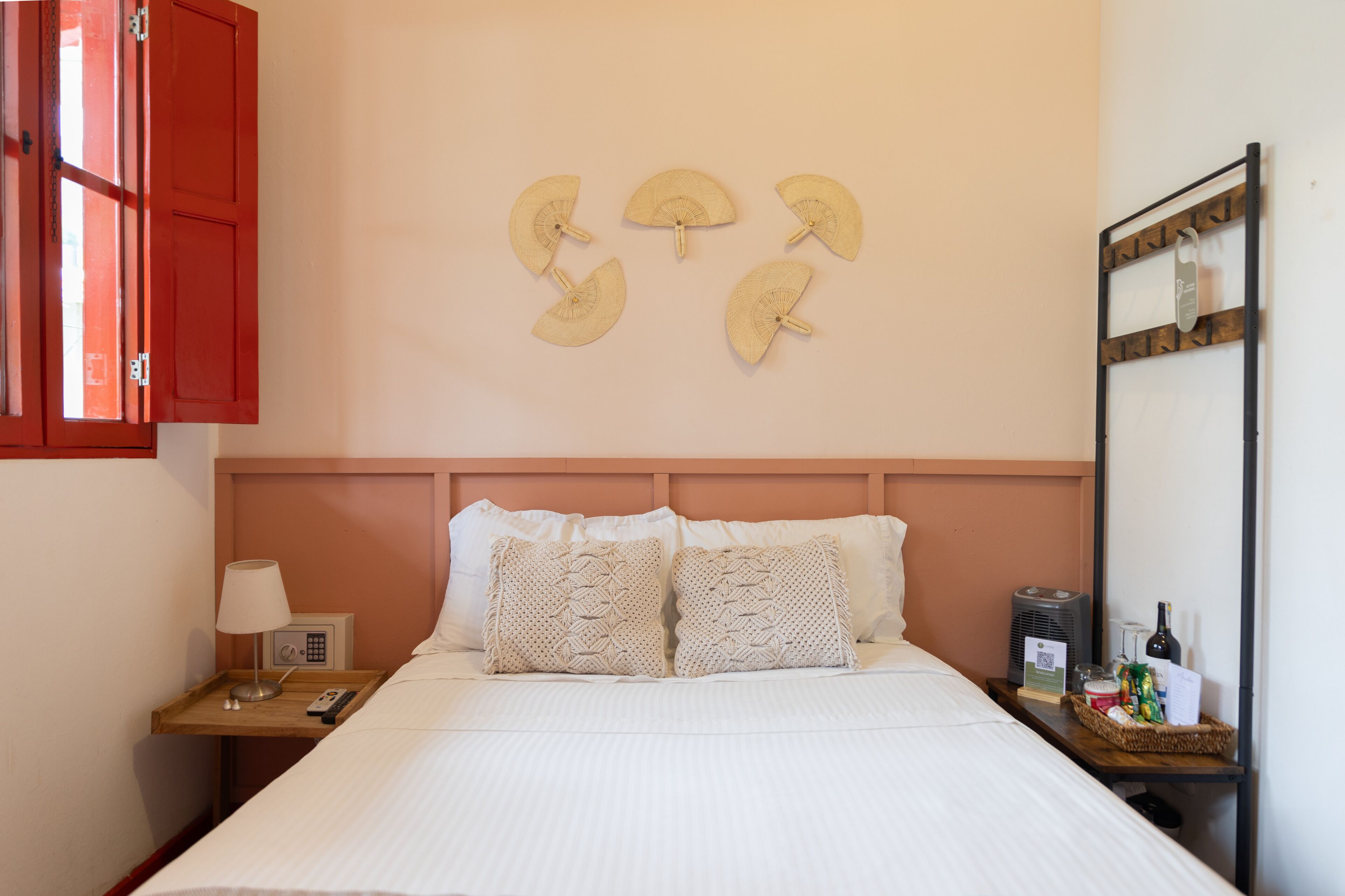 Una cama bien hecha con sábanas blancas, una mesita de noche de madera con una lámpara y una radio, y un estante en la pared con una botella y aperitivos.