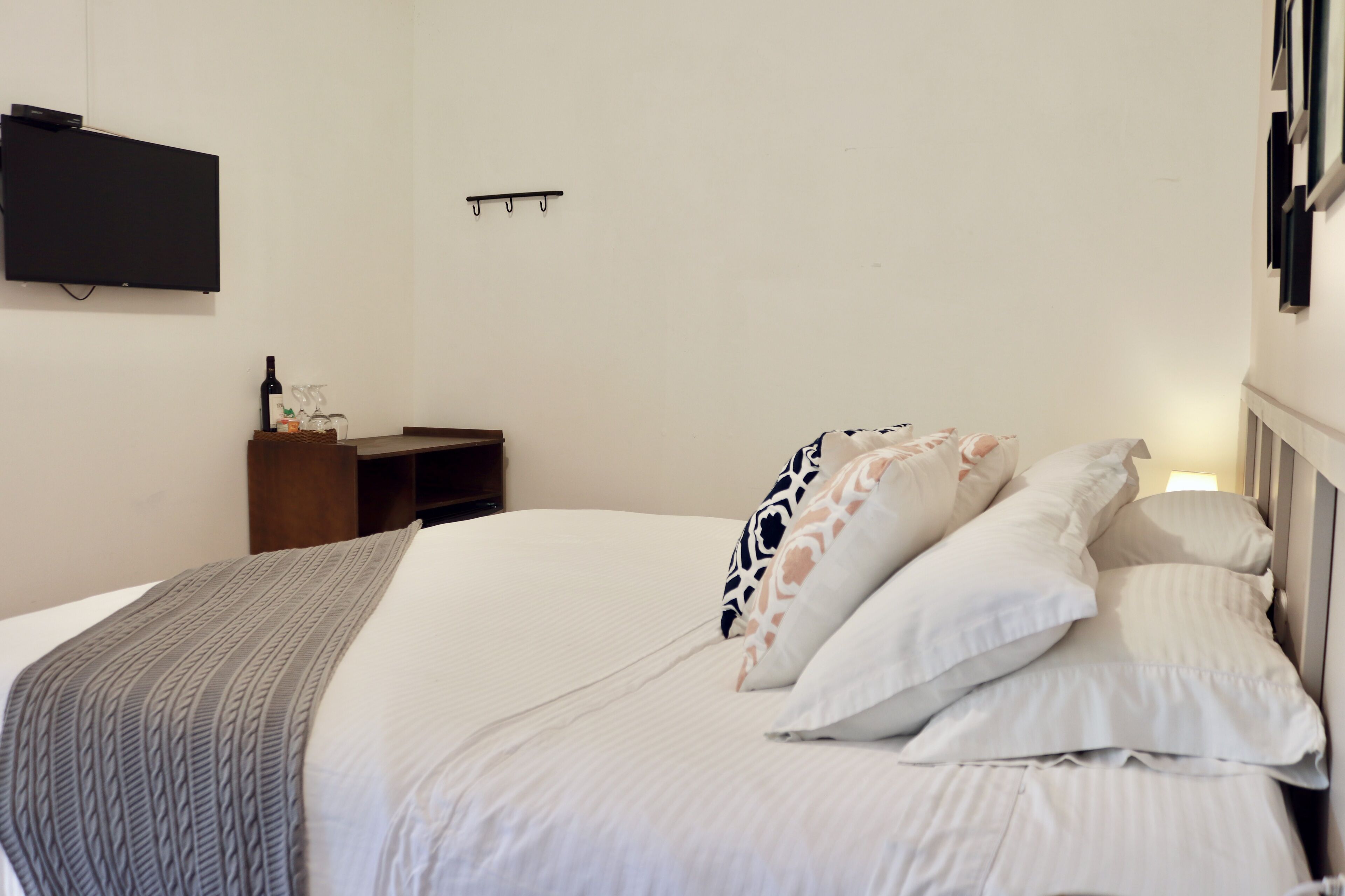 A neatly made bed with white and patterned pillows, a grey cable-knit throw, and a small wooden nightstand with a bottle and glasses.