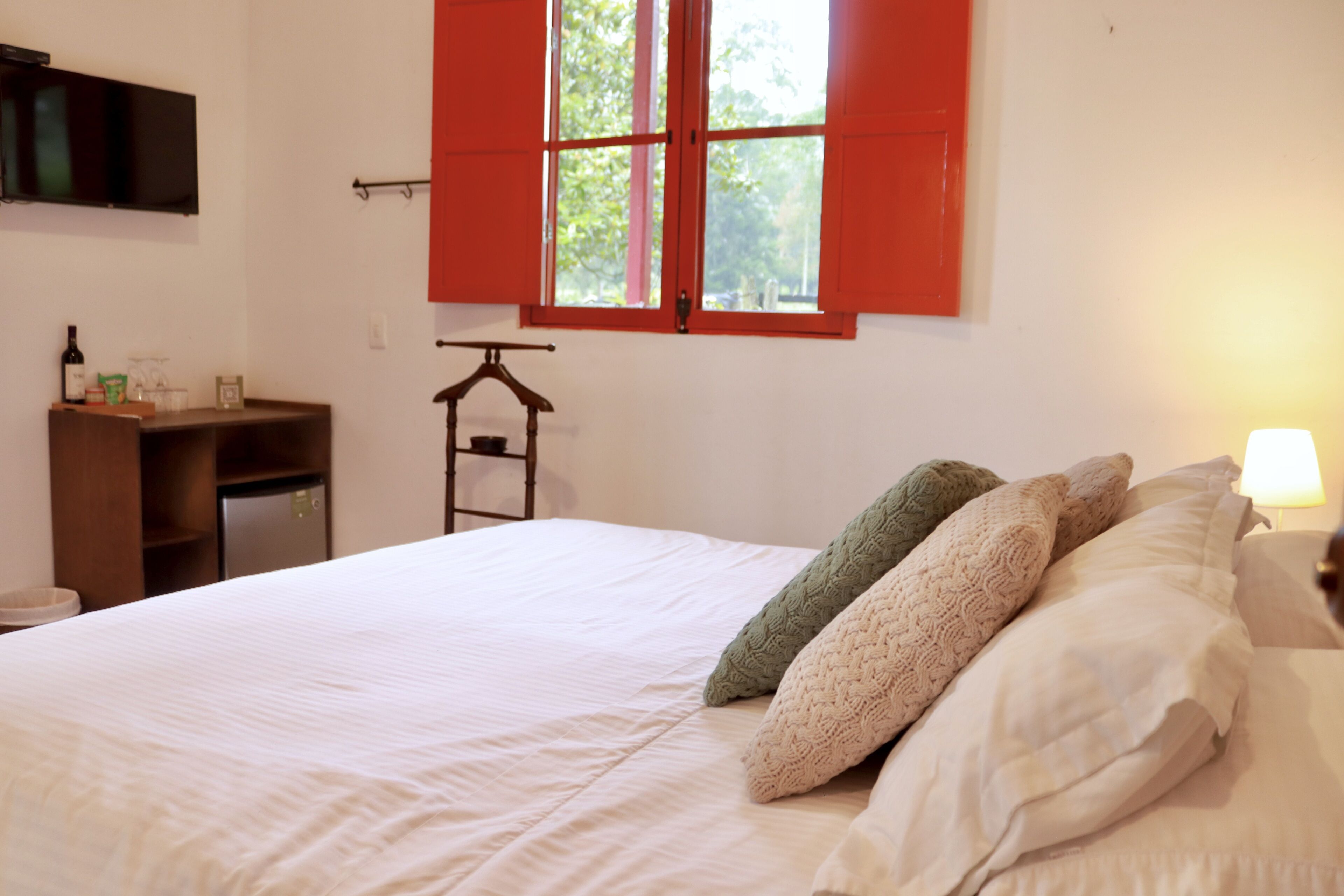 A bedroom with a bed, pillows, a nightstand, a lamp, a dresser, a TV, and a window with red shutters.