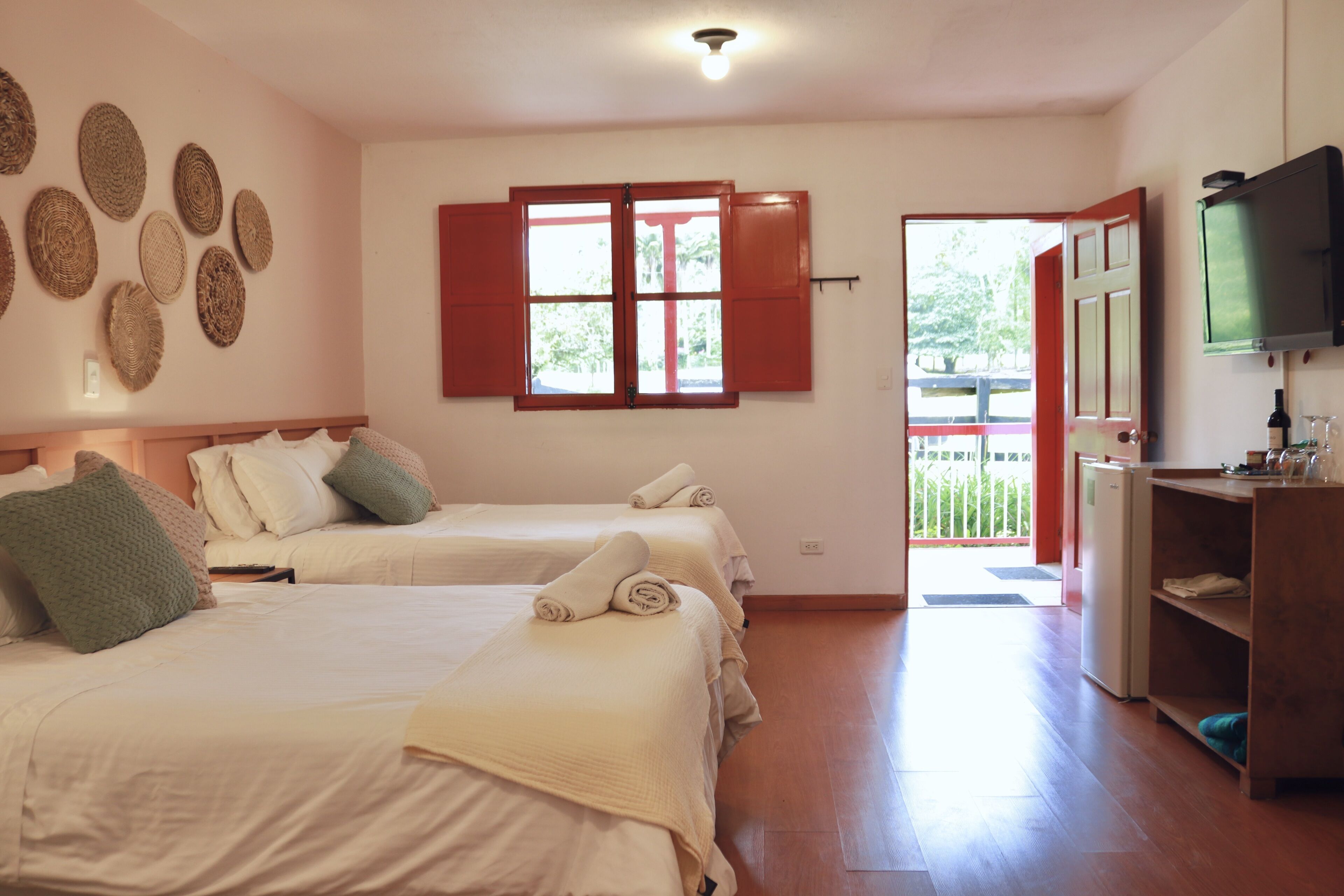 Habitación de hotel con dos camas, televisor, refrigerador y ventana con contraventanas rojas.