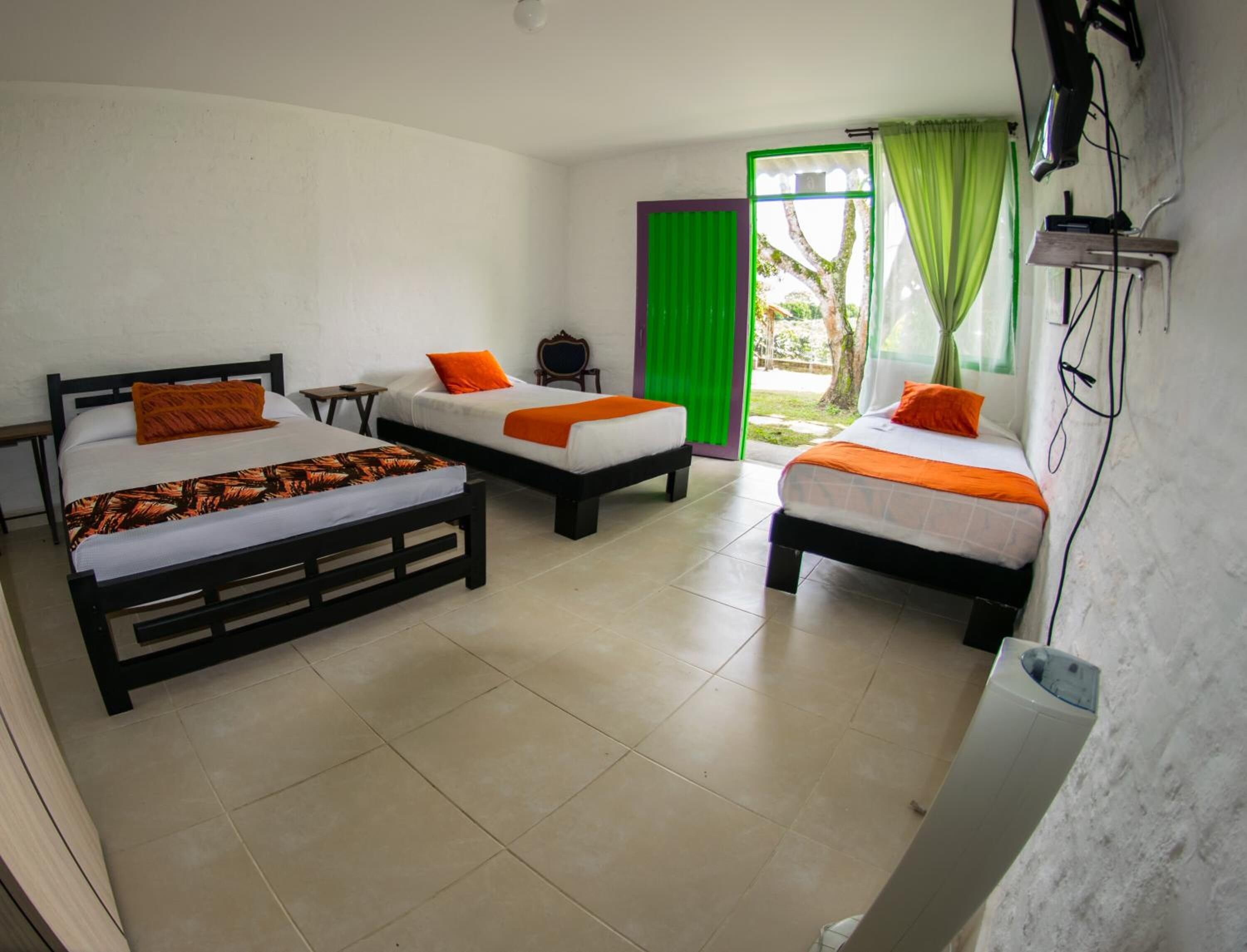 A room with three beds, each with an orange pillow and a patterned blanket. There is a small table and a chair, and a TV mounted on the wall.