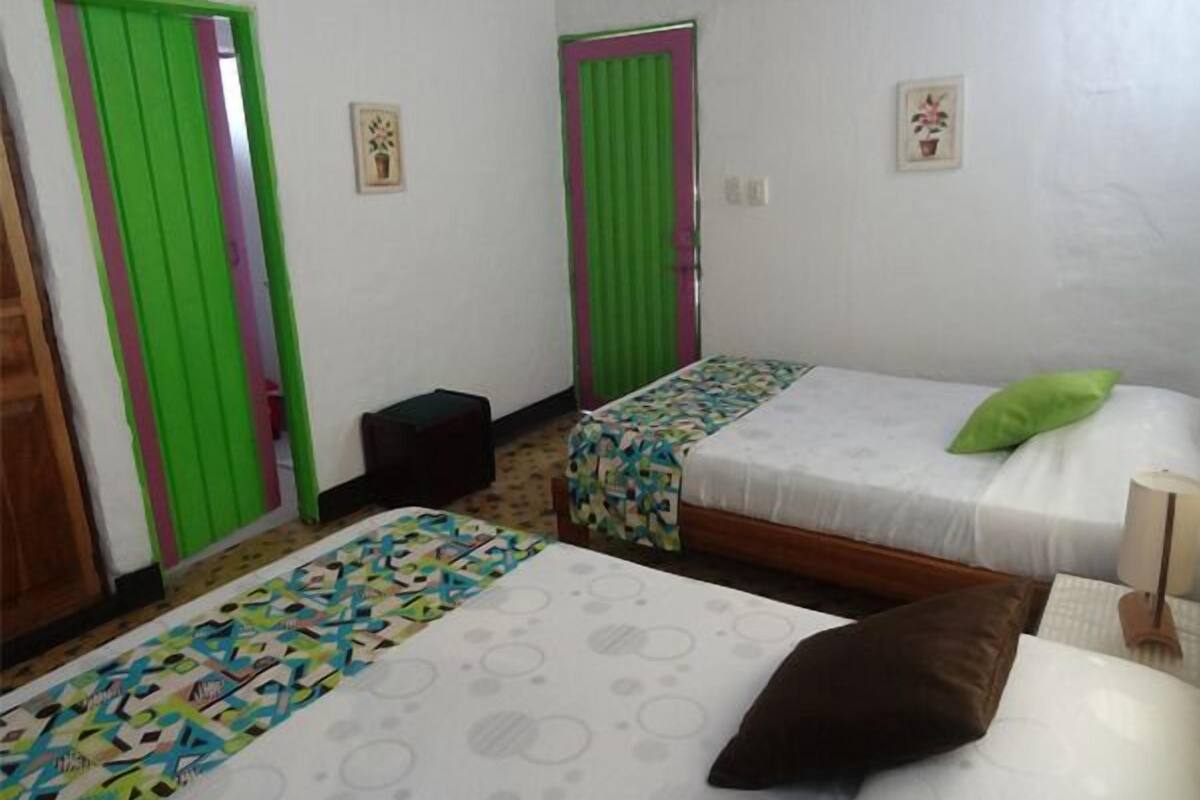 Ein Zimmer mit zwei Betten, jedes mit einer anderen Bettdecke. Grüne Türen und ein kleiner Hocker stehen in der Ecke.
