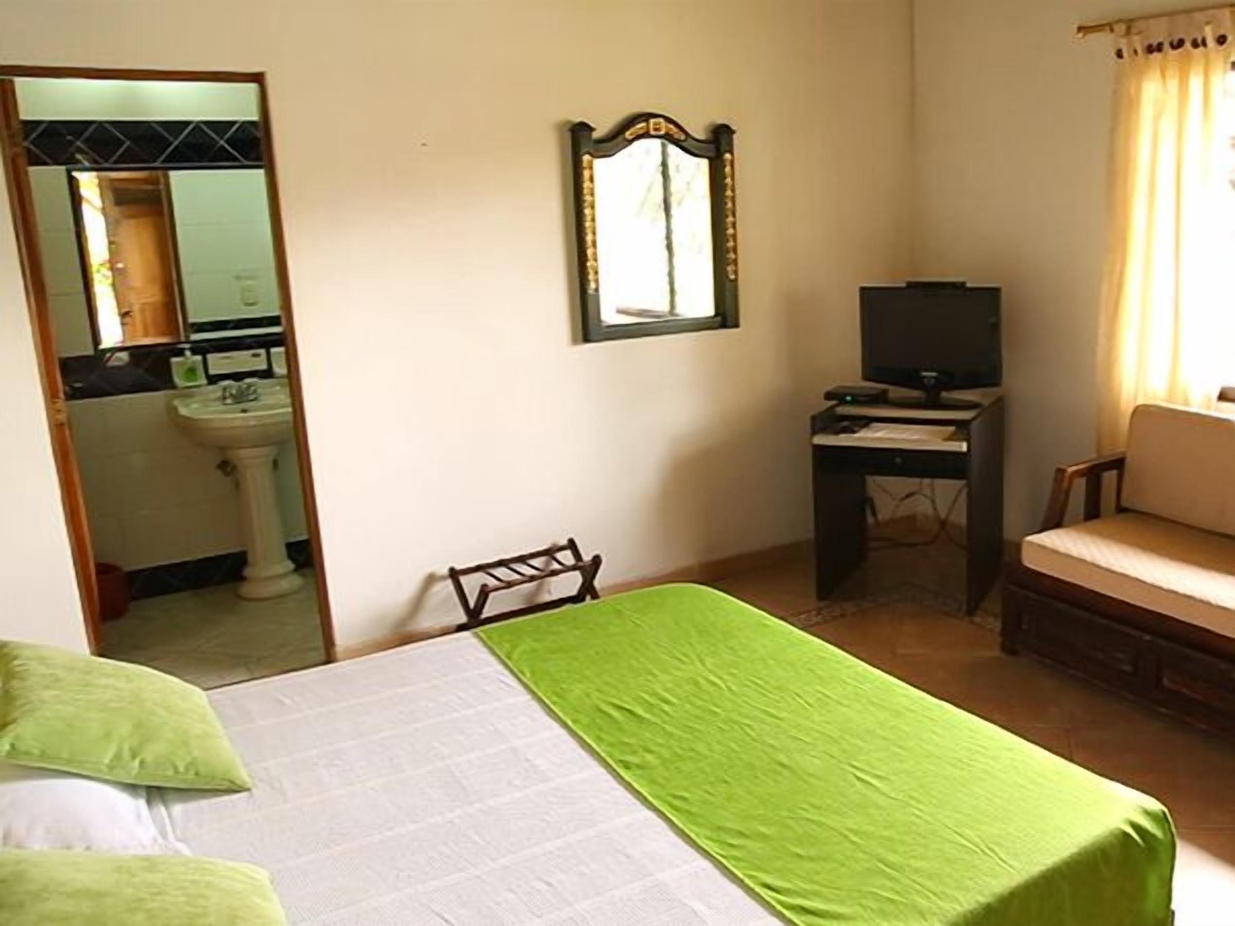 Un dormitorio con cama, escritorio con televisor, silla, sofá, espejo y baño visible a través de una puerta abierta.