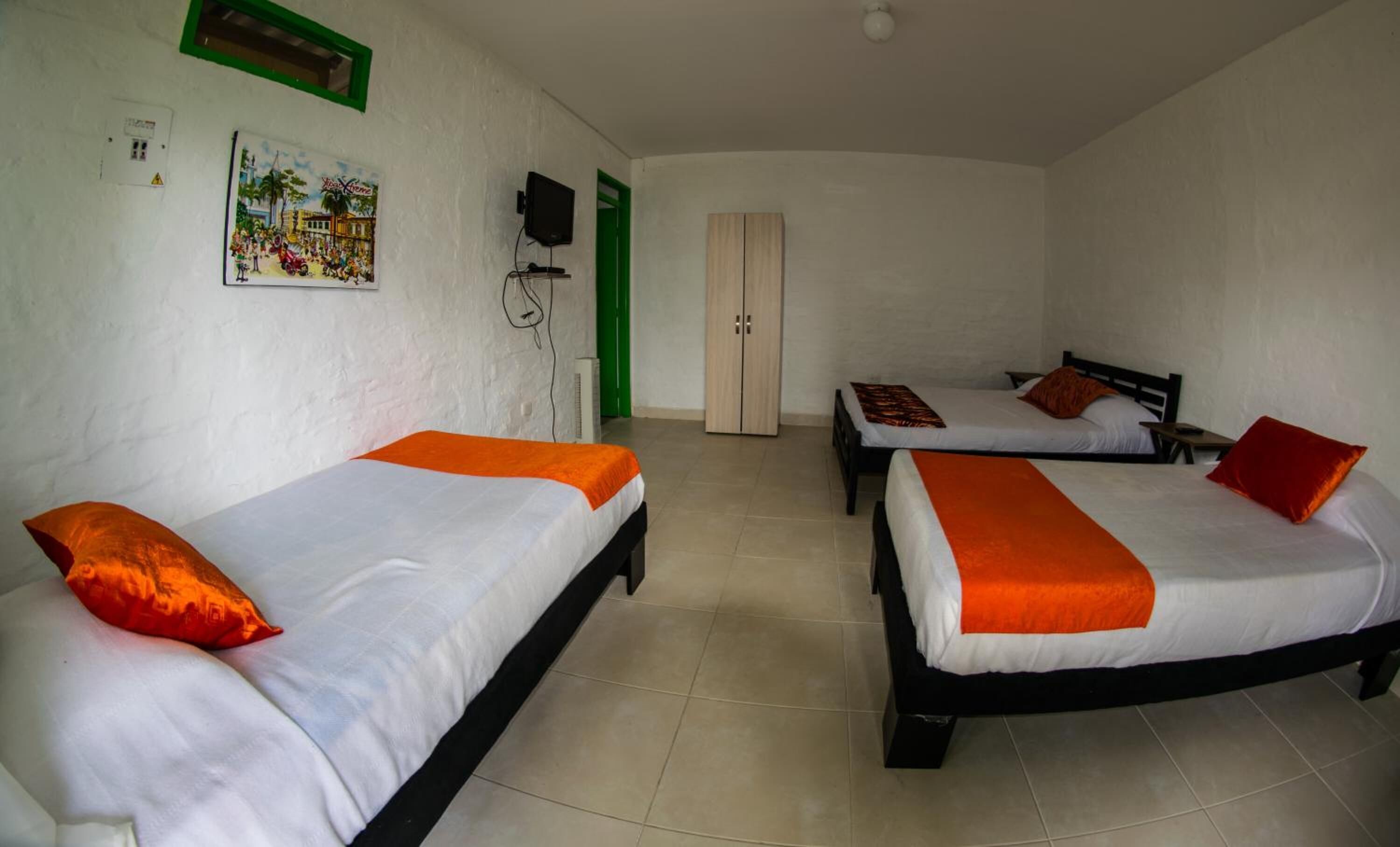 A room with three single beds, each with an orange blanket and pillow. There is a green door, a white wall with a framed picture, and a white ceiling with a light fixture.