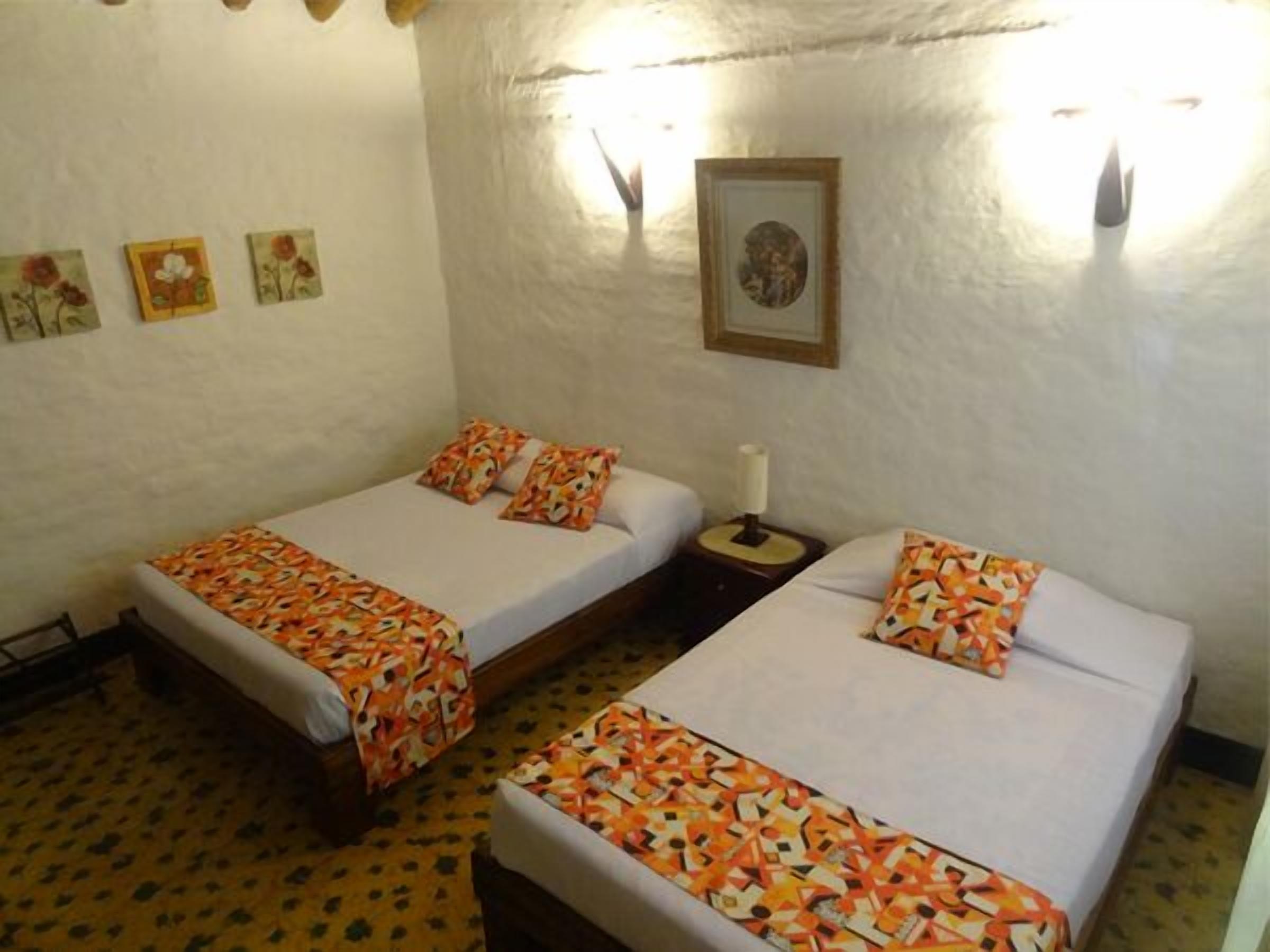 A room with two beds, each with patterned bedding and pillows. There are wall-mounted lights and a framed picture above the beds.