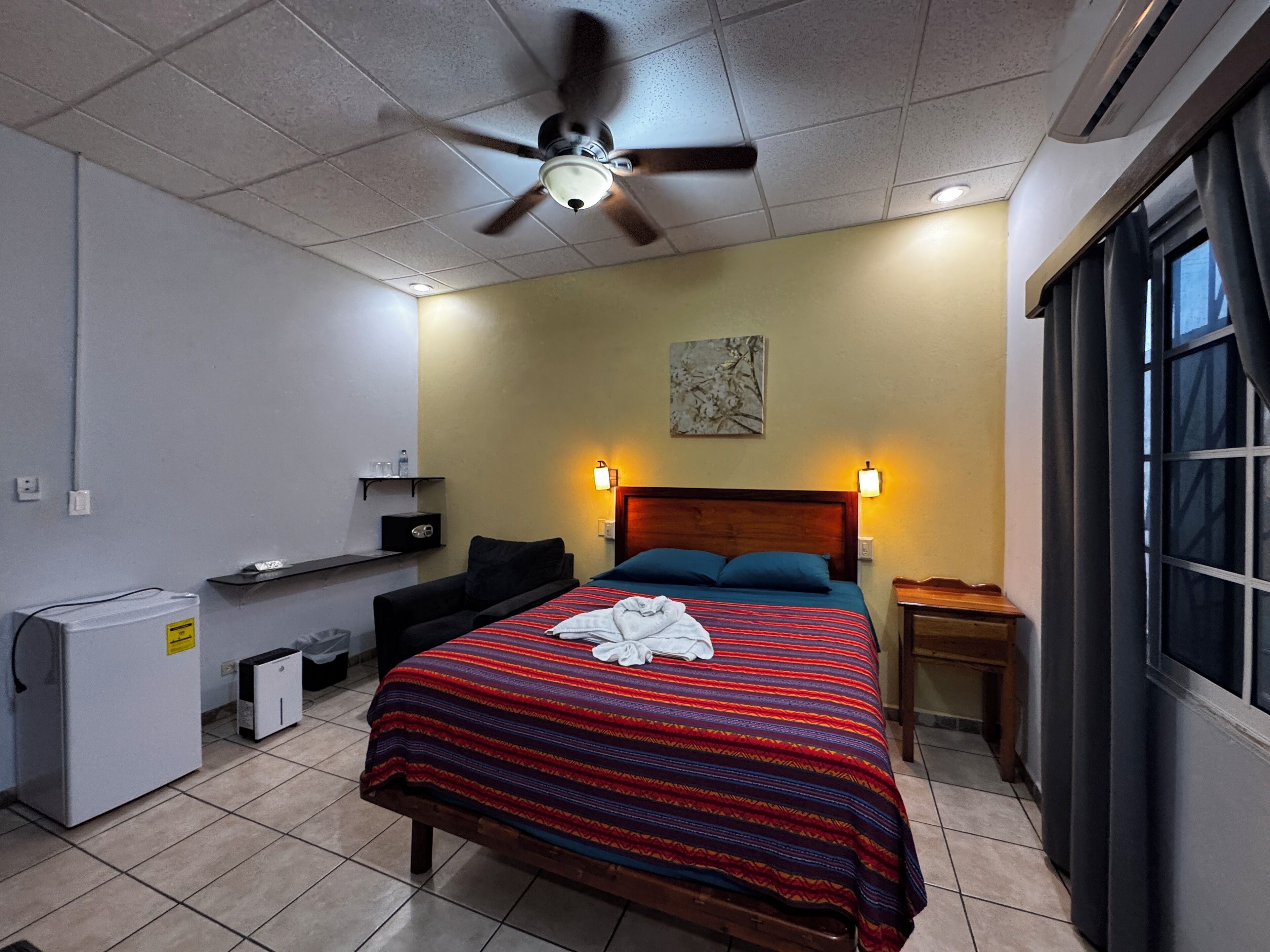 A hotel room with a bed, a ceiling fan, a nightstand, a window, and a small desk with a chair.