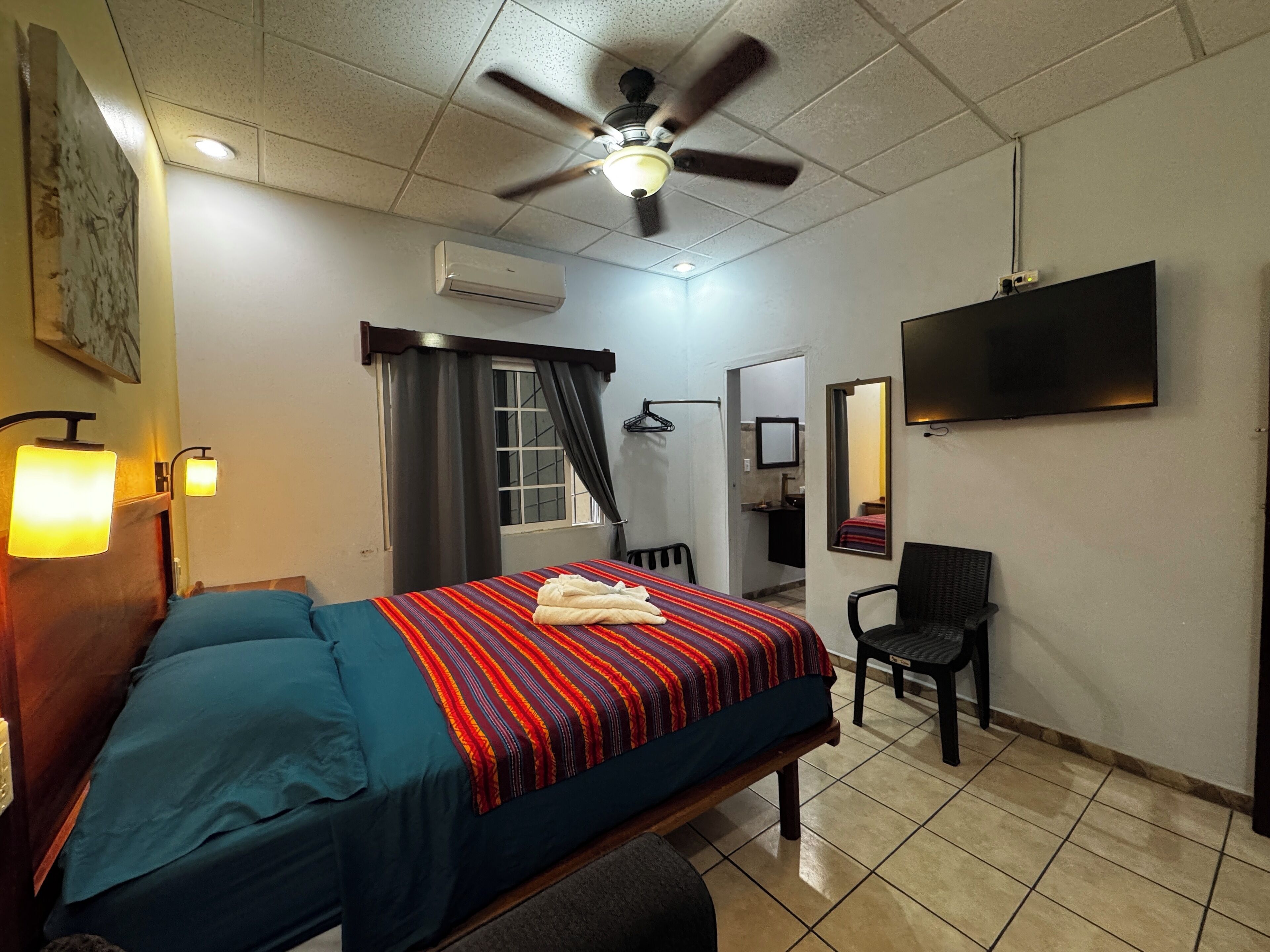 A hotel room with a bed, a ceiling fan, an air conditioning unit, a television, a mirror, and a bathroom visible through an open door.