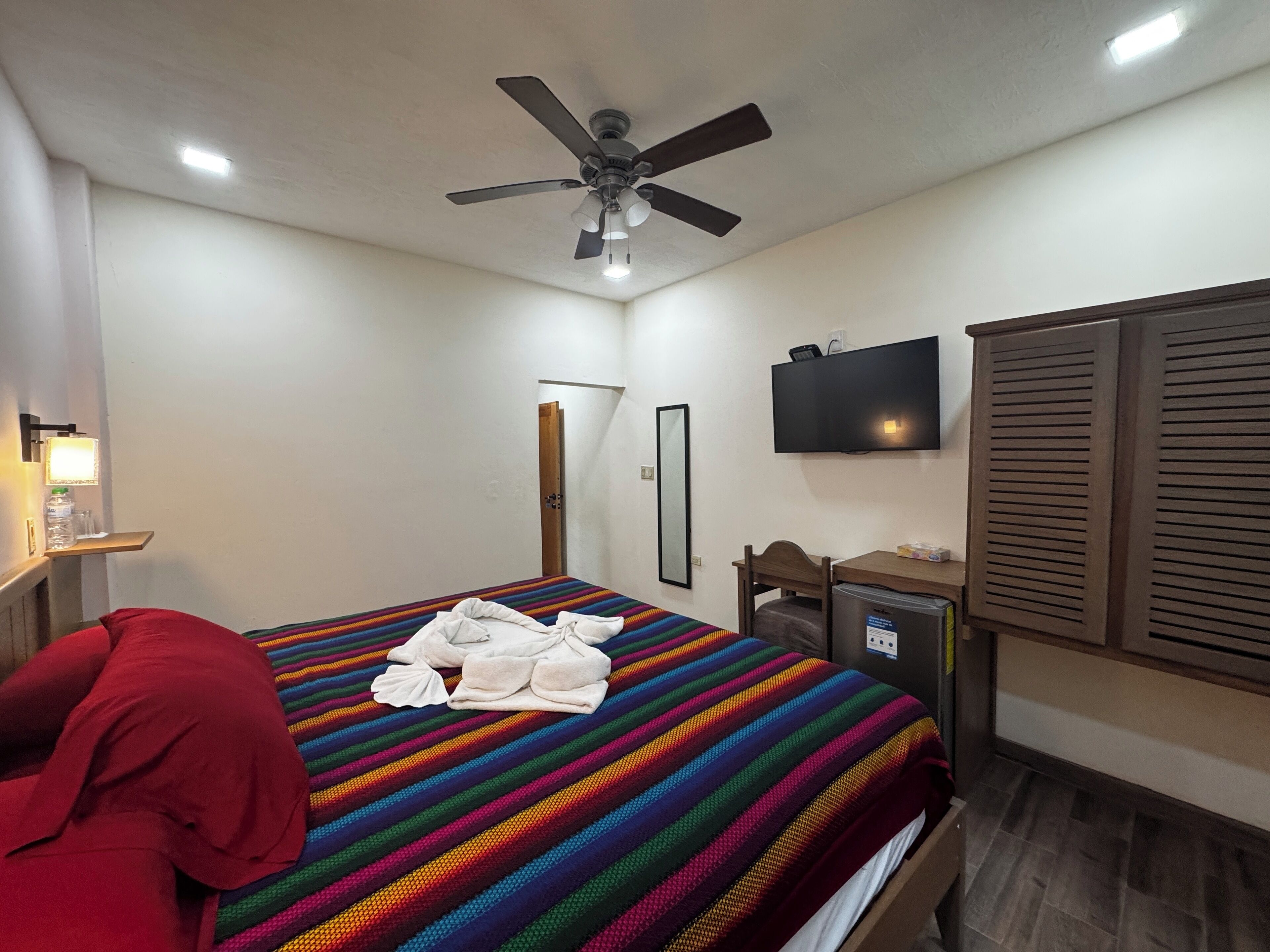 A hotel room with a bed, a ceiling fan, a TV, a desk, and a mirror.