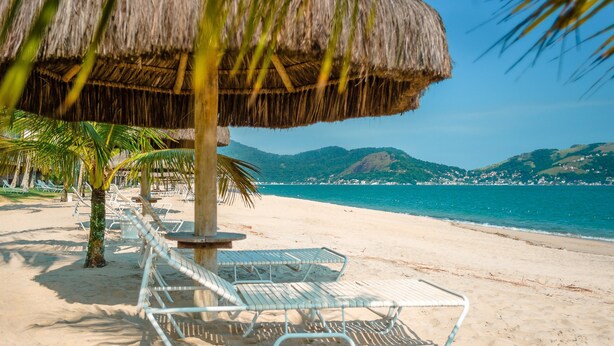 A beach with thatched umbrellas, palm trees, and metal lounge chairs. - Portobello Resort & Safári (Mangaratiba)