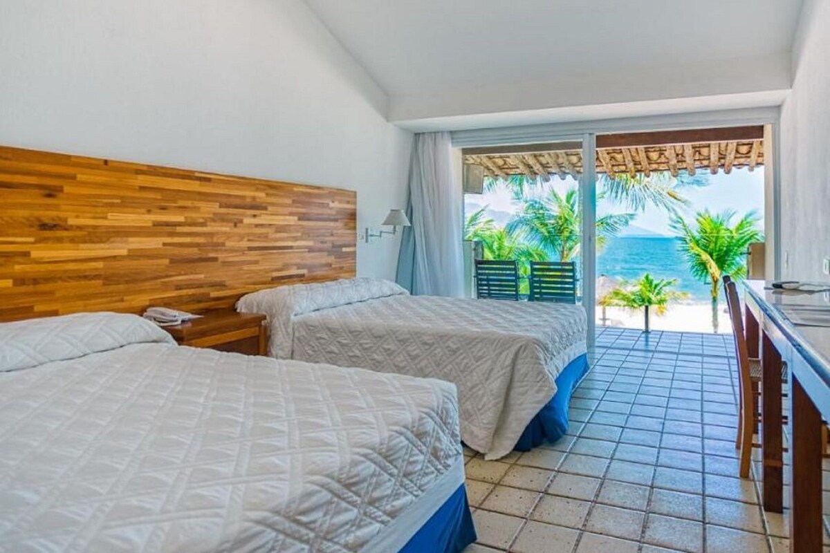 Ein Hotelzimmer mit zwei Betten, einem Holzkopfteil, Meerblick und einem Balkon mit Tisch und Stühlen.