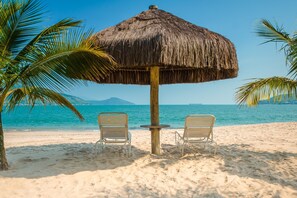 Escena de playa con una sombrilla de paja, dos sillas de playa y una palmera.