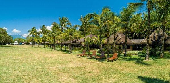 A resort with thatched-roof huts, palm trees, and sun loungers on a grassy lawn.