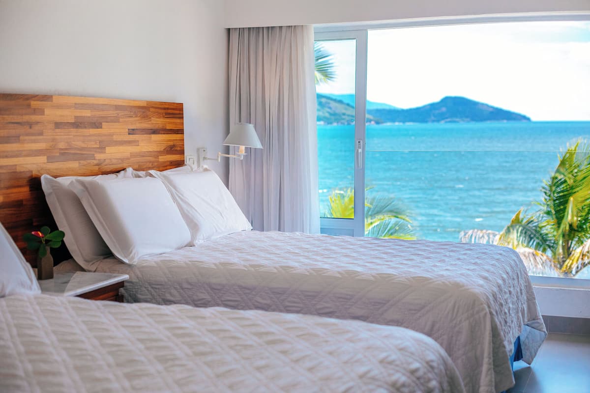 Een hotelkamer met twee bedden, een houten hoofdbord, een nachtkastje met een lamp en uitzicht op zee en bergen.