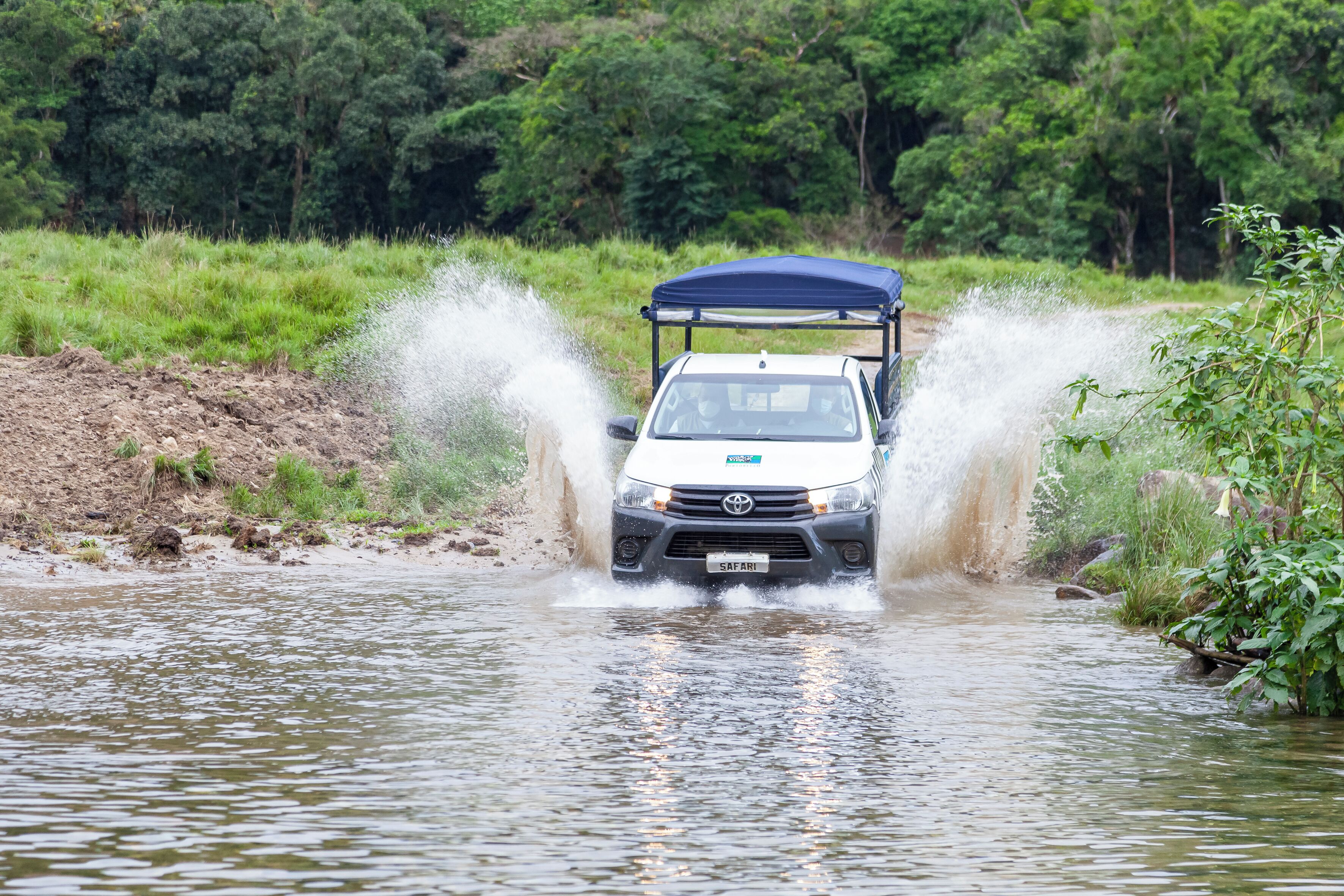 A vehicle driving through a shallow water body, surrounded by greenery.