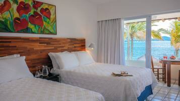 A hotel room with two beds, a wooden headboard, a painting, a balcony with a table and chairs, and a view of the sea.