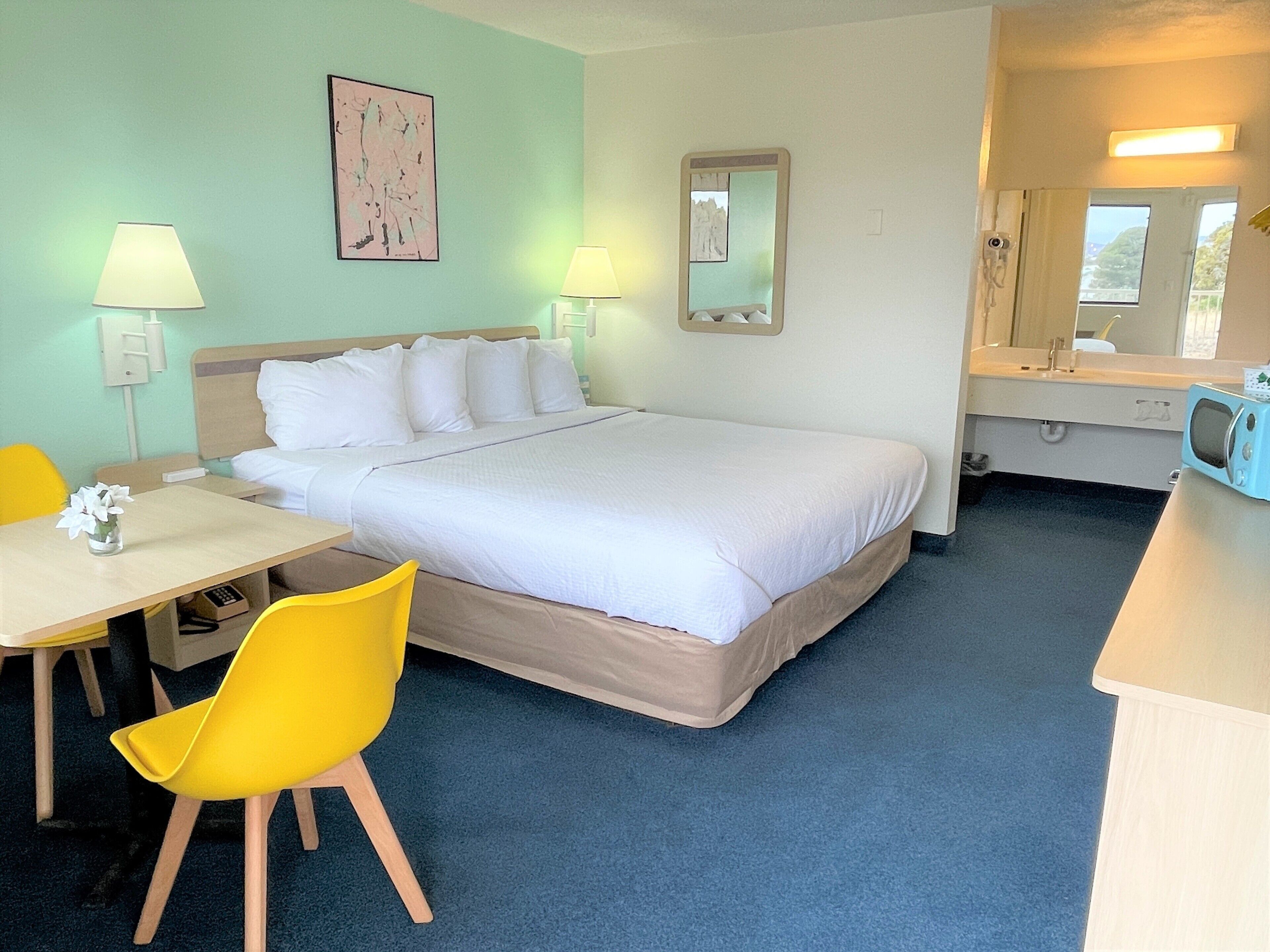 A hotel room with a bed, a desk with a chair, a lamp, a mirror, and a microwave.