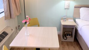 Habitación de hotel con una cama, una mesita de noche con lámpara, una pequeña mesa con un florero, una silla y un calefactor.
