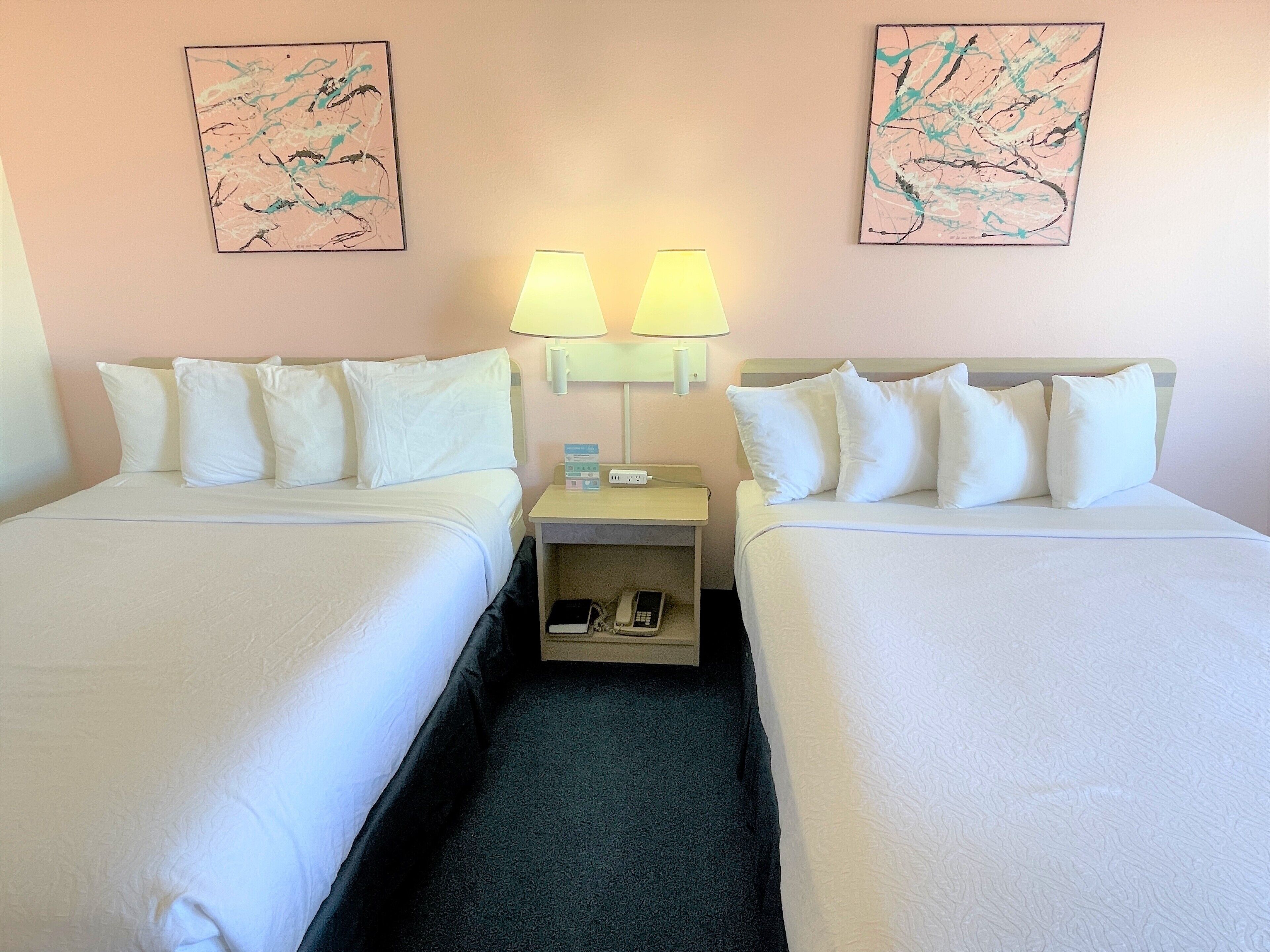 A hotel room with two beds, each with white linens and pillows, a nightstand with a lamp, and two abstract paintings on the walls.
