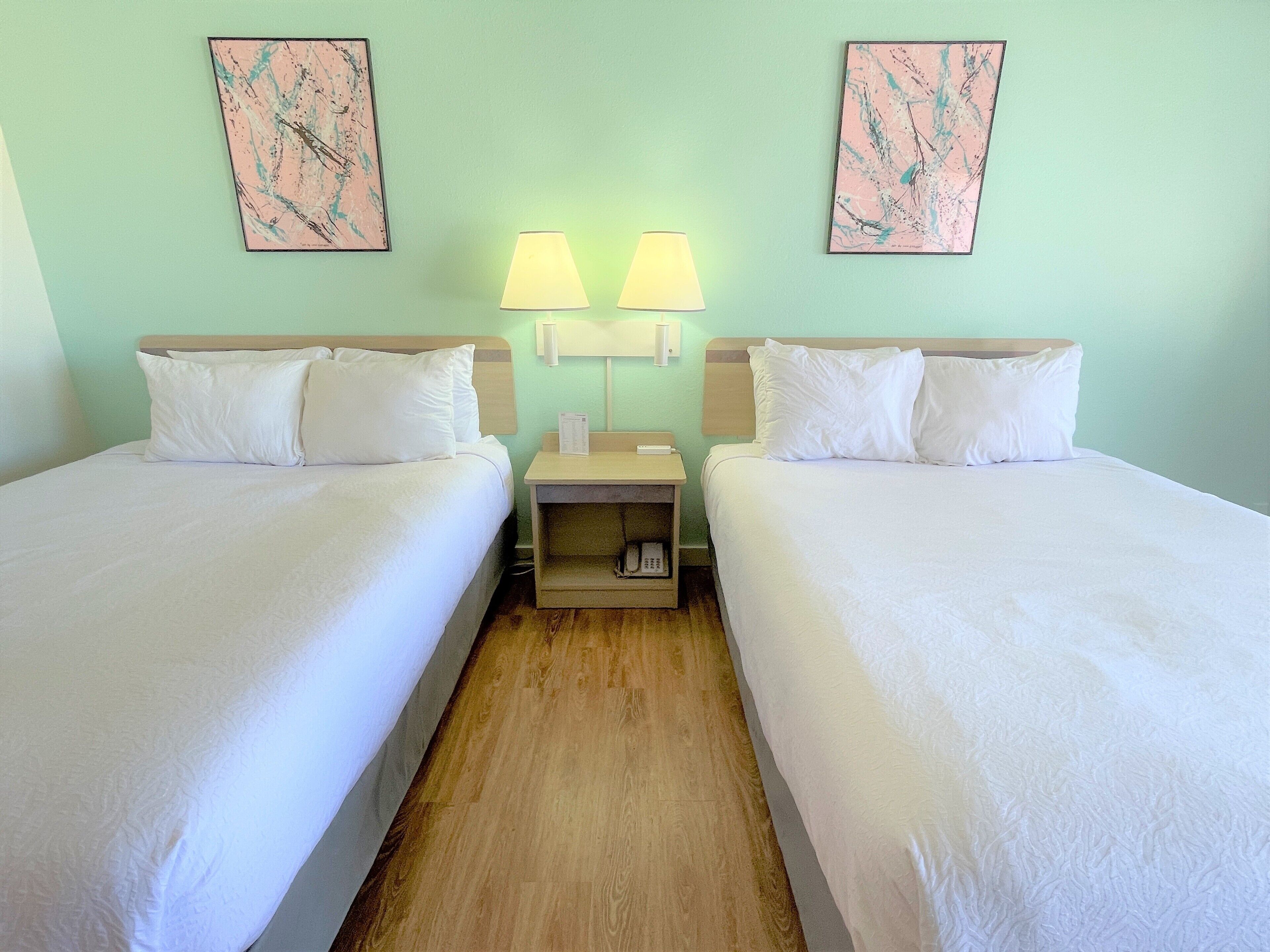 Une chambre d’hôtel avec deux lits simples, une table de chevet en bois avec une lampe, et deux tableaux abstraux encadrés accrochés au mur.