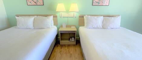 Une chambre d’hôtel avec deux lits simples, une table de chevet en bois avec une lampe, et deux tableaux abstraux encadrés accrochés au mur.