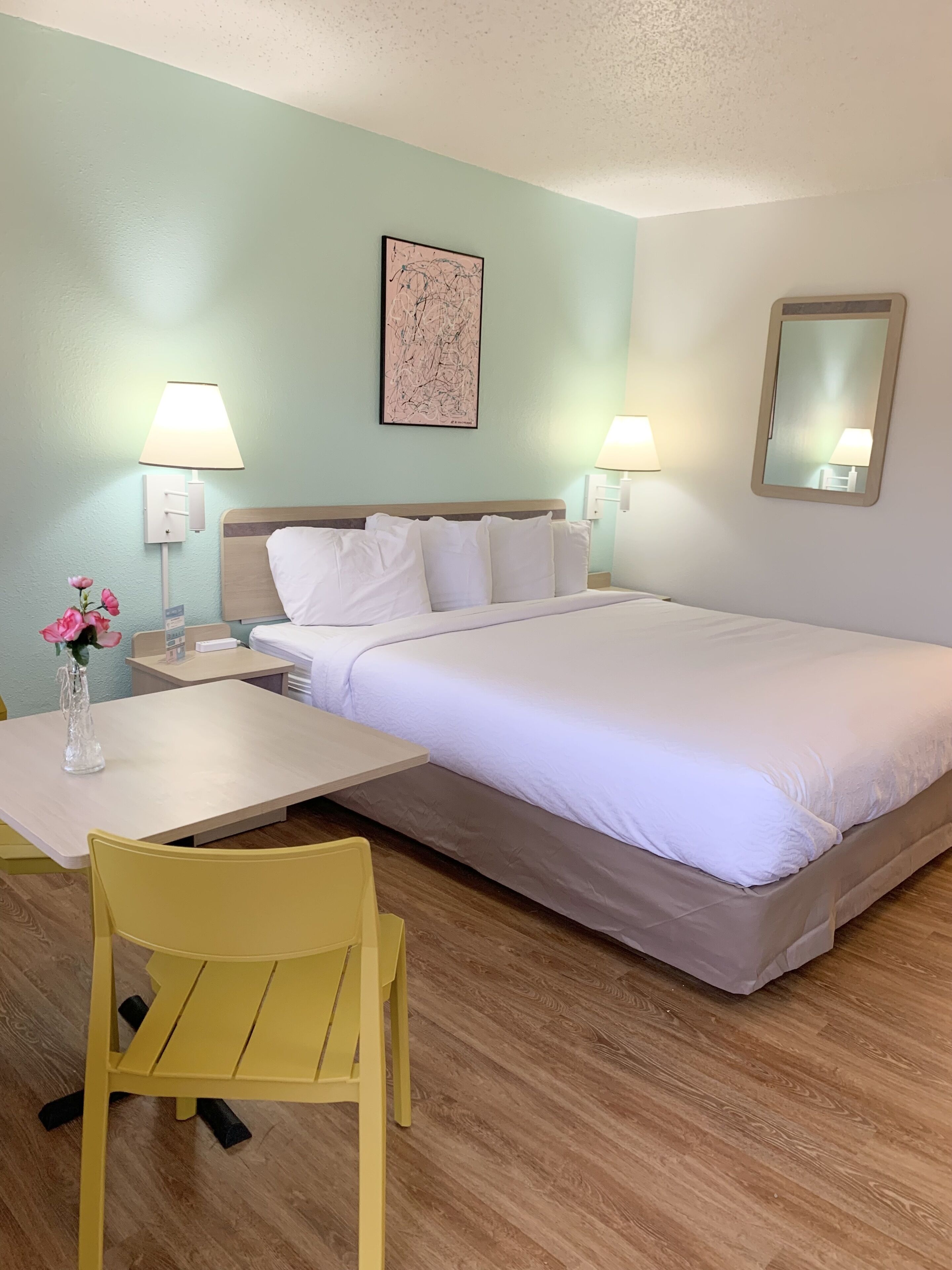 A hotel room with a bed, a desk, a chair, a nightstand, a lamp, a vase with flowers, and a wall art piece.