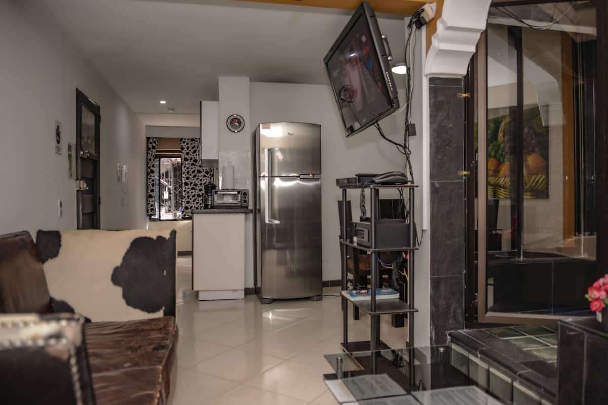 A modern living room with a sofa, a flat-screen TV mounted on the wall, a kitchen area with a refrigerator, and a dining table.