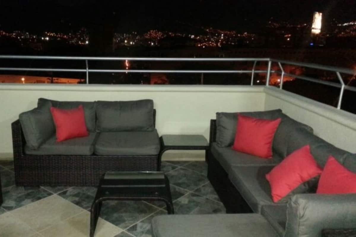 A rooftop terrace with two grey sofas and red cushions, a small table, and a wicker chair.